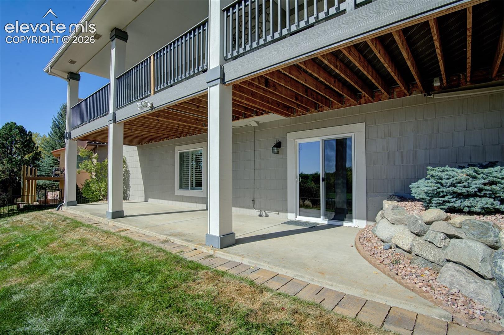 Covered concrete patio featuring structural support columns, dual-level railed balcony, sliding glass door, and natural stone landscaping