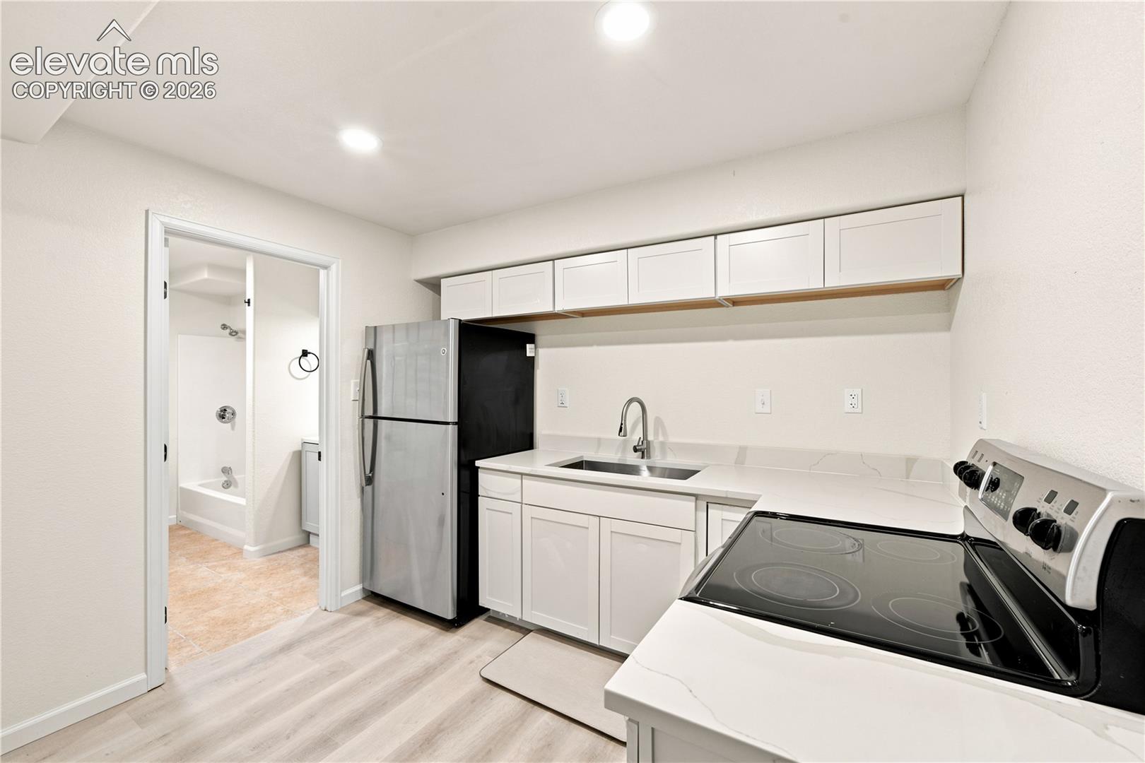 Compact kitchen area featuring wood-finish flooring, white cabinetry, integrated stainless steel sink with gooseneck faucet, full-size refrigerator, and electric range