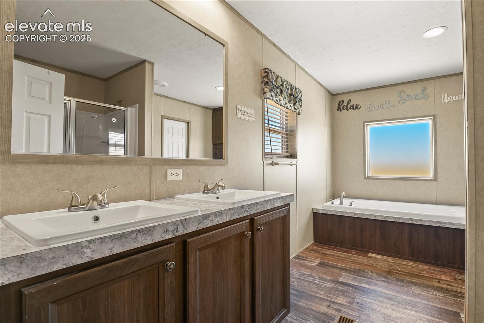 Bathroom featuring a dual-sink vanity with light-toned countertops, recessed lighting, wood-finish flooring, and a large soaking tub with a wood-panel surround