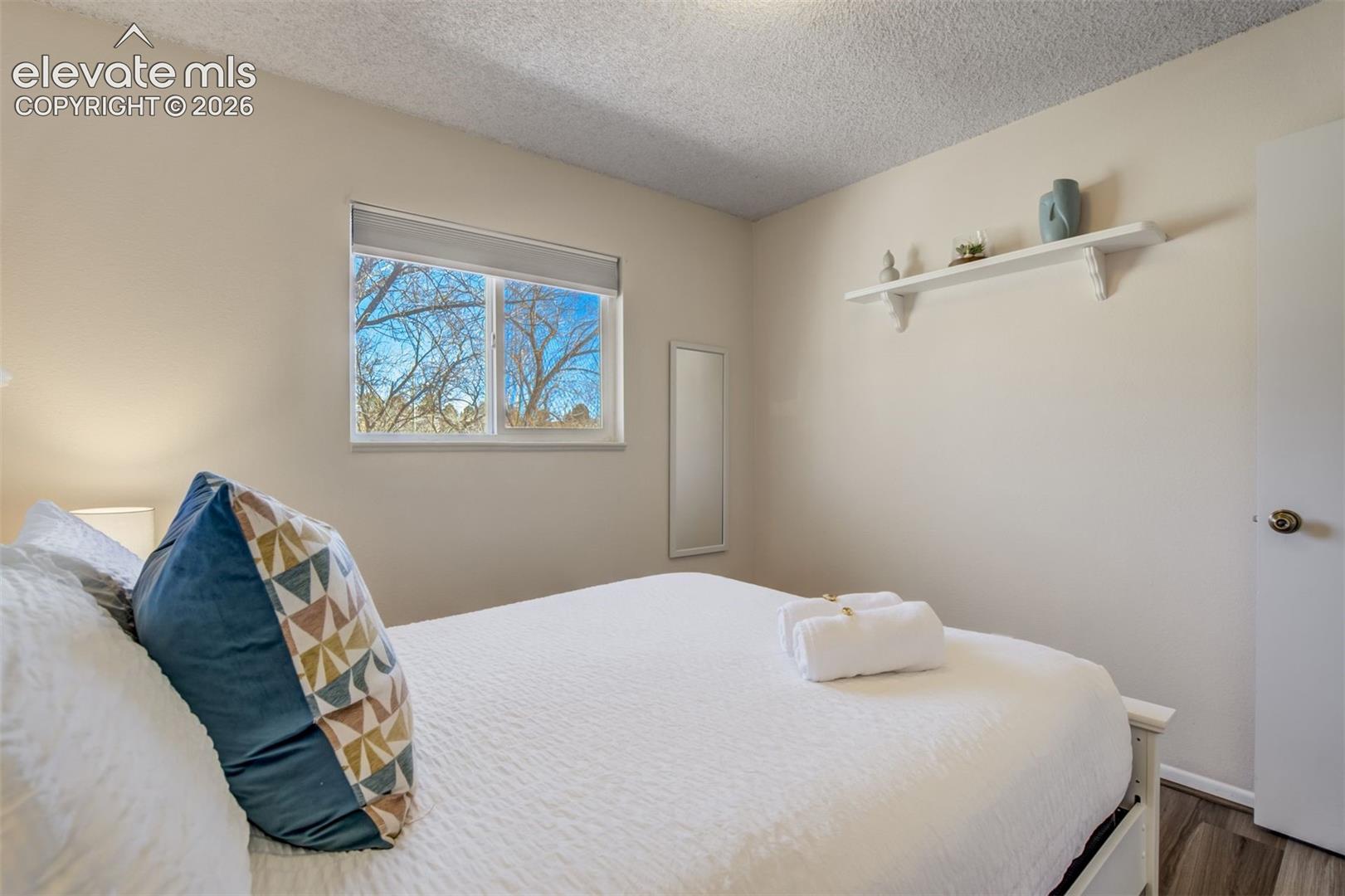 Window with a roller shade, textured ceiling, light-colored walls, wood-finish flooring, and a wall-mounted shelf