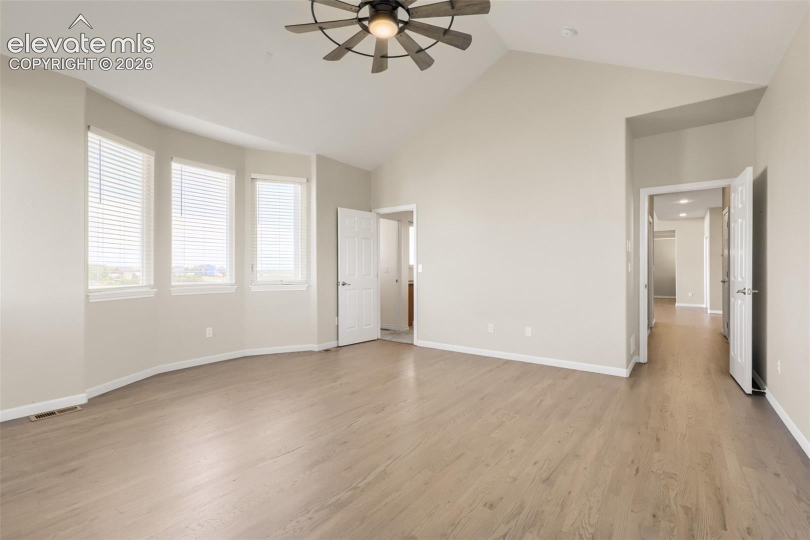 Spacious and serene main floor primary suite featuring dramatic vaulted ceilings and a statement windmill ceiling fan. Beautiful hardwood floors sweep through the room, while a sun-drenched bay of large windows floods the space with natural light.