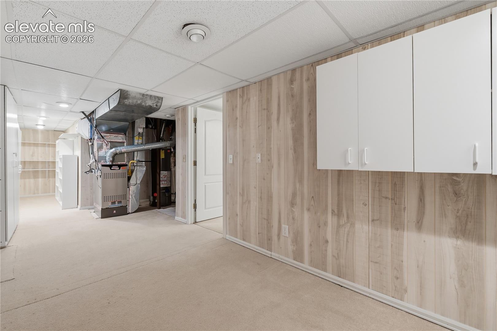 Utility area featuring wall-mounted white cabinetry, wood-finish paneling, drop ceiling, and carpeted flooring