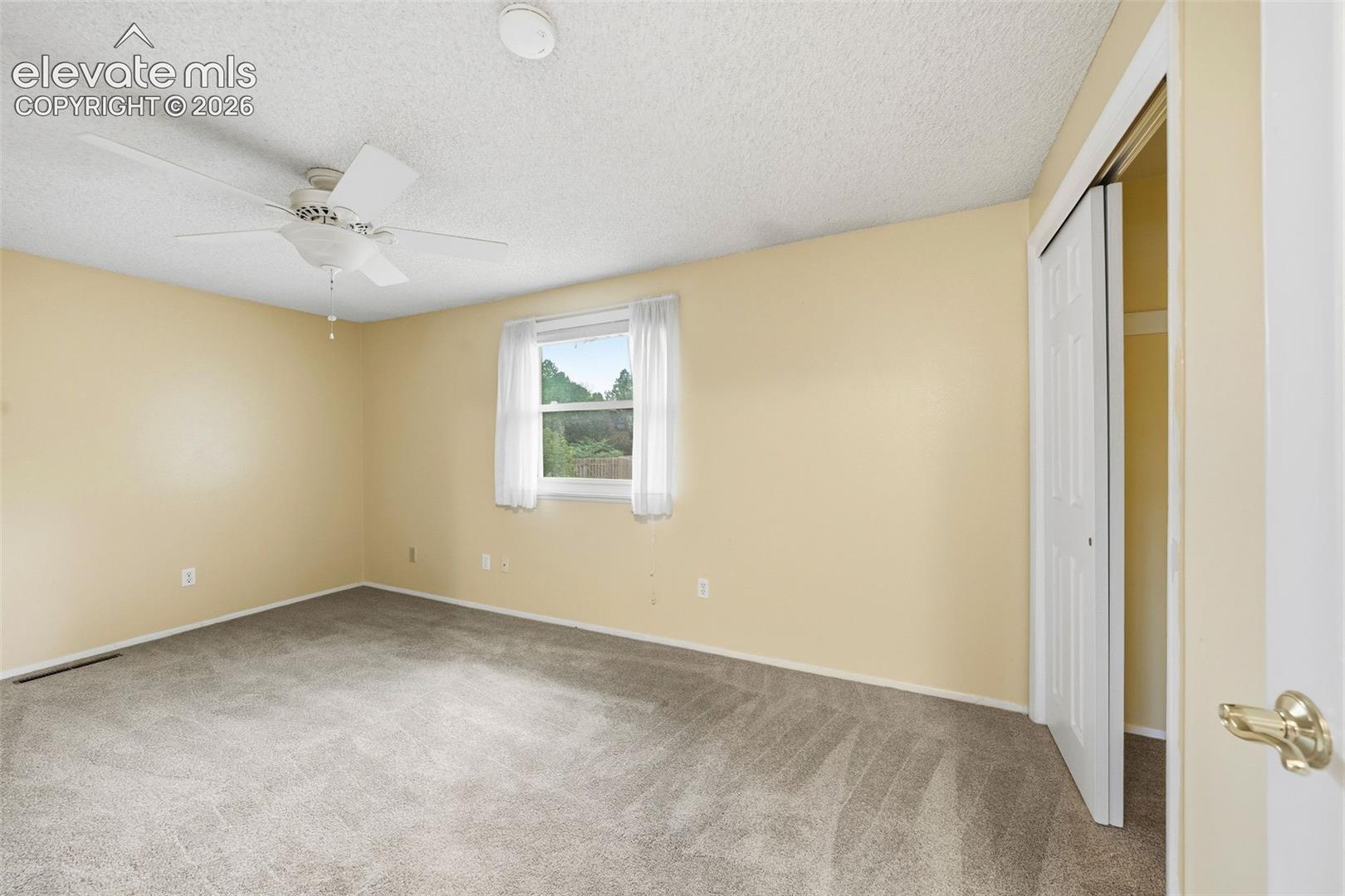 Neutral-toned carpeting, ceiling fan with light fixture, dual-panel closet door, and single-pane window
