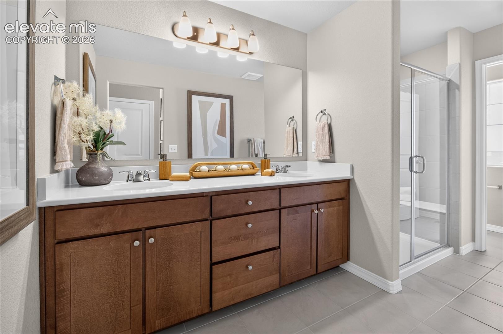 Double vanity with wood-finish cabinetry, solid-surface countertop, dual undermount sinks, and a full-width mirror with four-light fixture, featuring a glass-enclosed shower