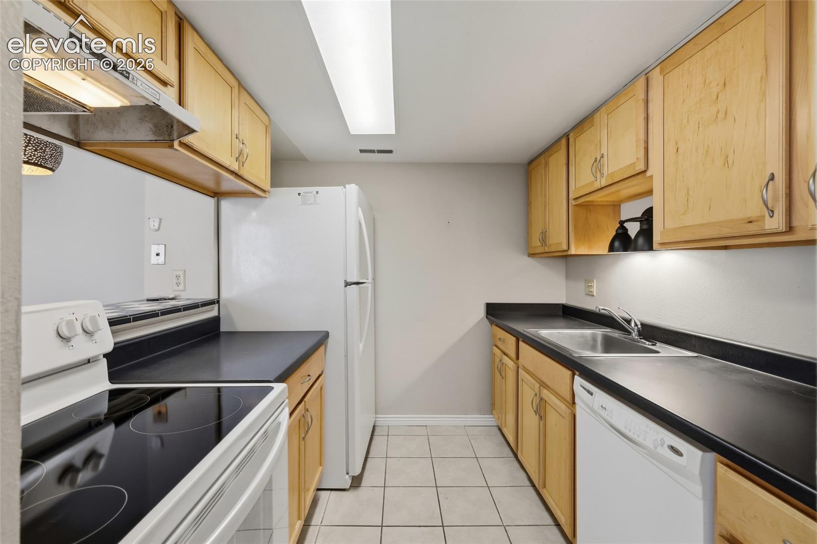 Galley kitchen featuring light wood-finish cabinetry, dark solid-surface countertops, tile flooring, and white appliance suite