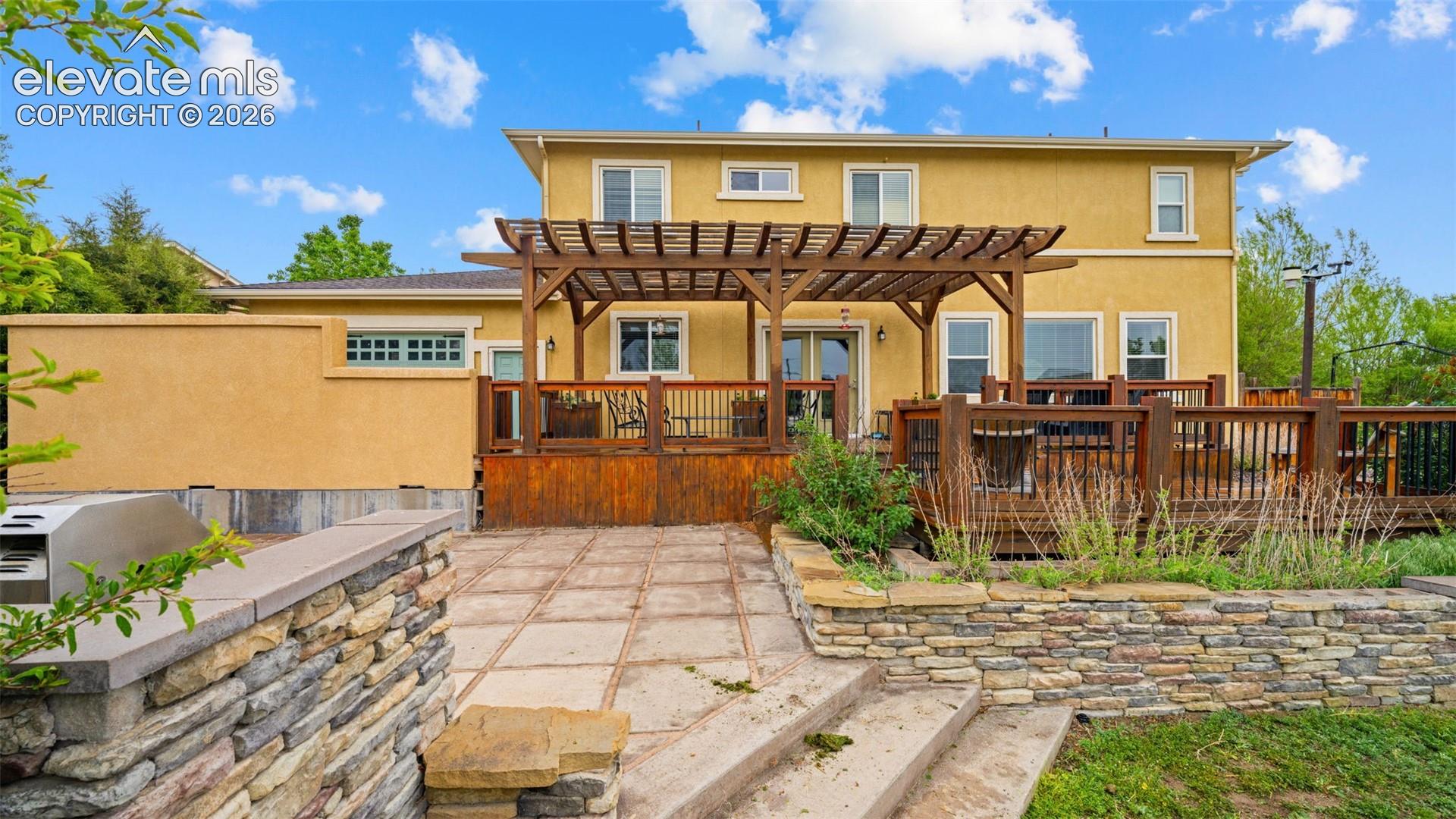 Multi-level backyard featuring a spacious wood deck with pergola, stone retaining walls, and a built-in outdoor grill station with stone finish