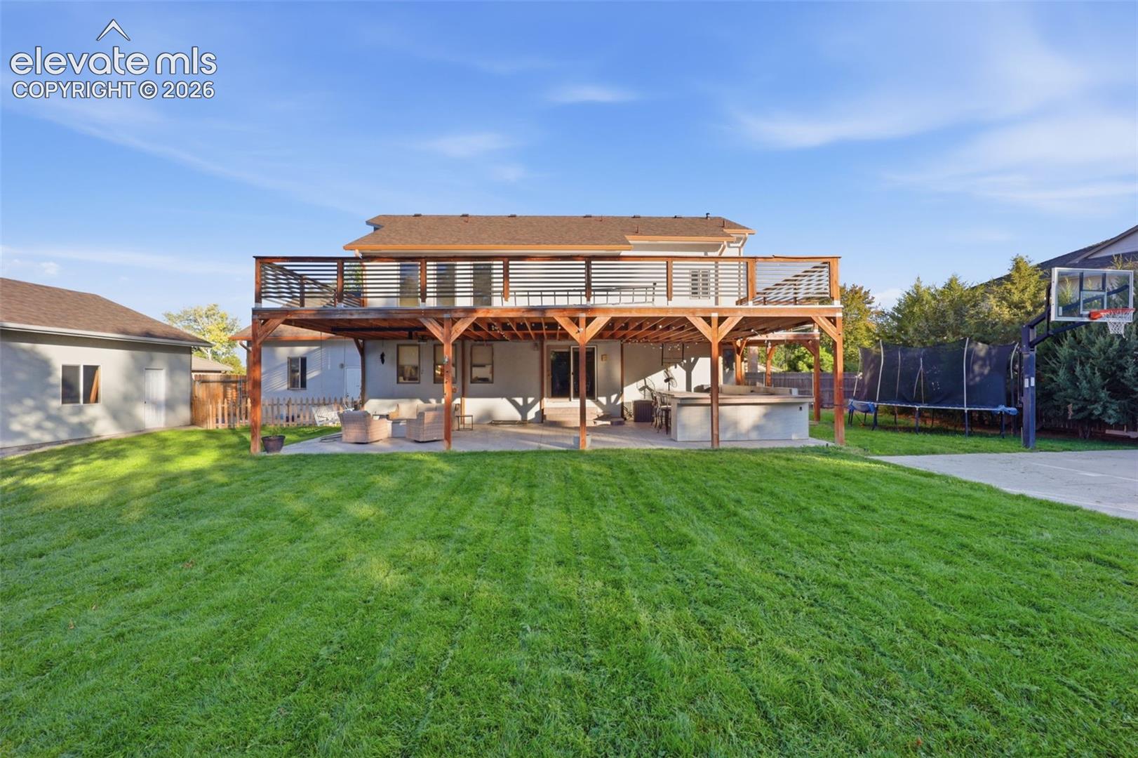 Expansive backyard featuring a manicured lawn, multi-level wooden deck with horizontal slat railings, a ground-level concrete patio, and a dedicated concrete sports court