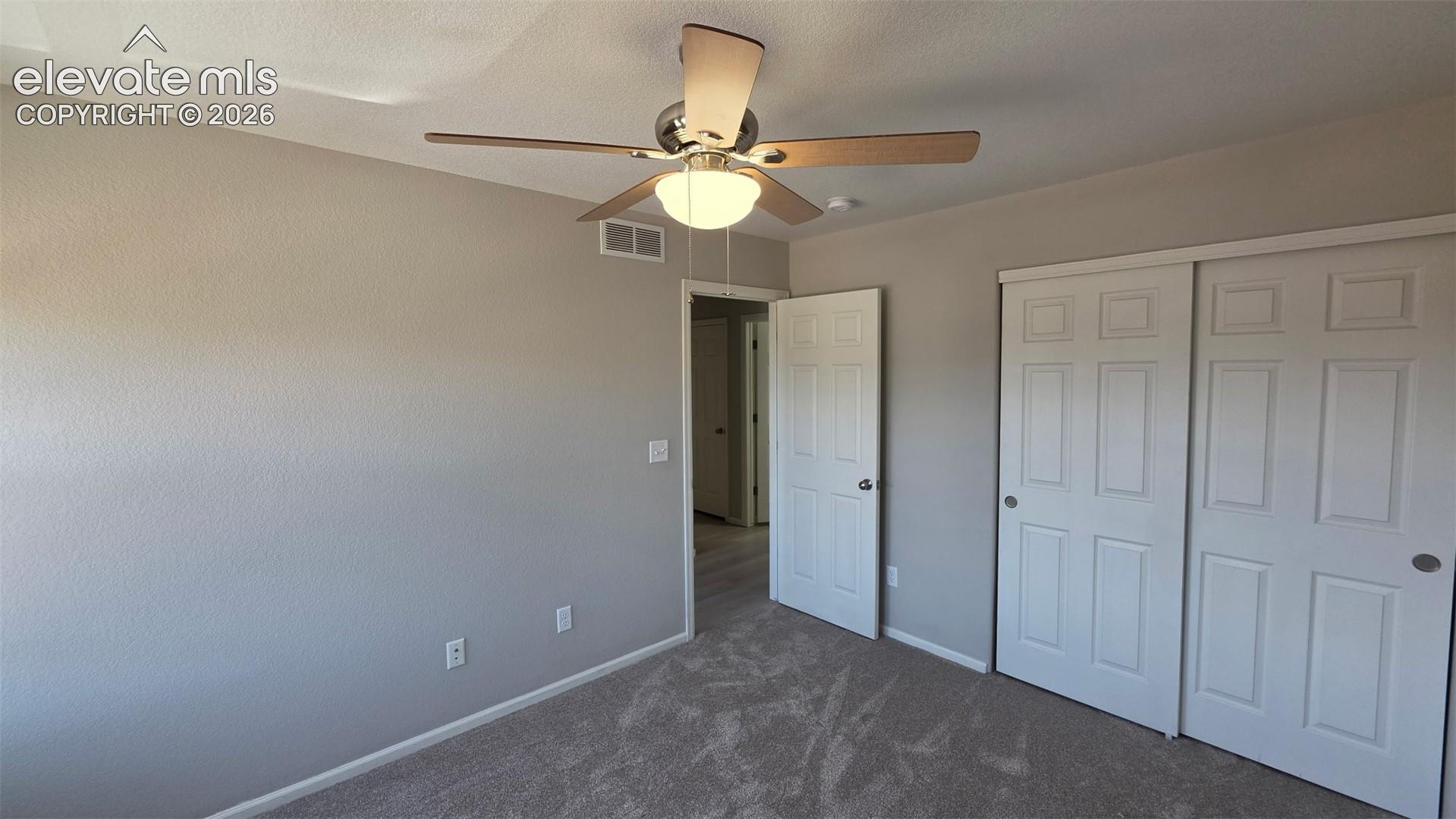 Neutral-toned room featuring textured wall finish, plush carpeting, a multi-blade ceiling fan with integrated lighting, and white six-panel closet doors