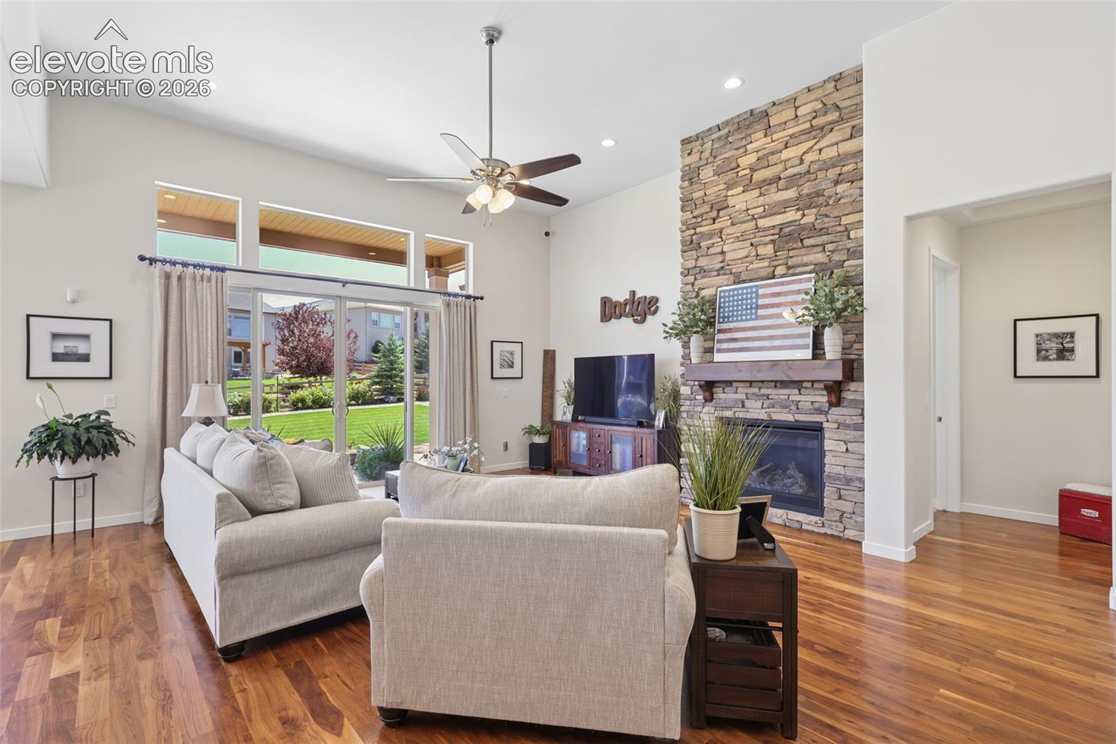 Vaulted ceiling with a ceiling fan, stone-clad fireplace, wood mantel, glass sliding door, and wood-finish flooring