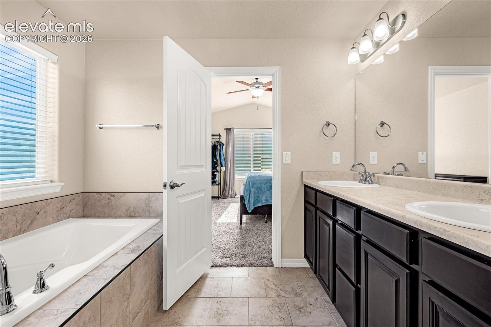 Soaking tub with tile surround, dual vanity with undermount sinks, wood-finish cabinetry, and tile flooring