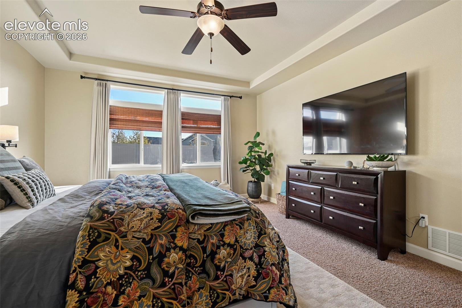 Tray ceiling with a ceiling fan, wall-mounted sconce lighting, and multiple windows with blinds