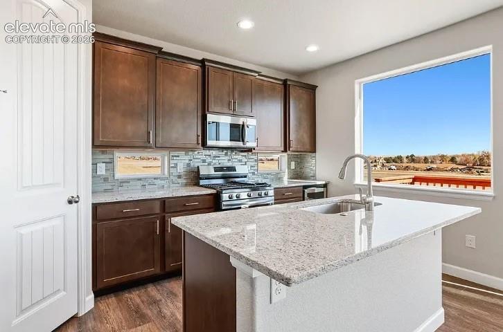 Kitchen featuring wood cabinetry, stone-surface countertops, center island with undermount sink, multi-tone tile backsplash, and wood-finish flooring