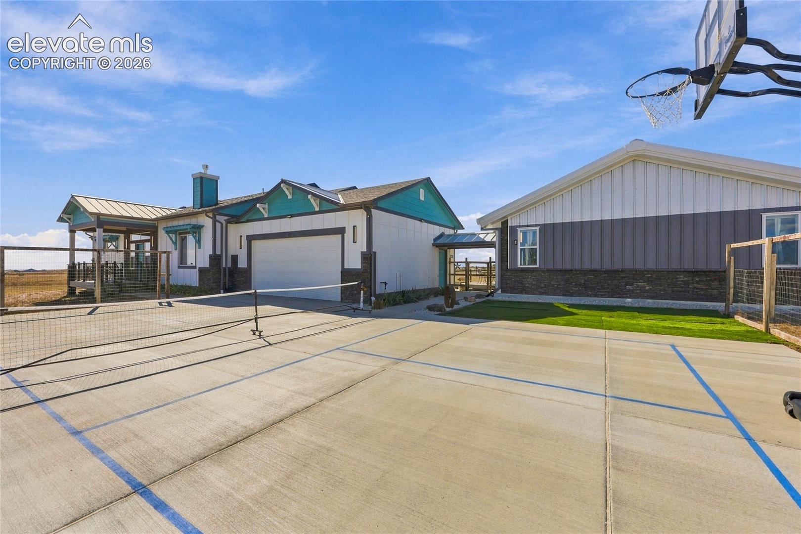 Concrete sports court featuring a painted boundary grid, basketball hoop, and tennis net, situated on a lot with an exterior metal-sided structure and attached multi-car garage