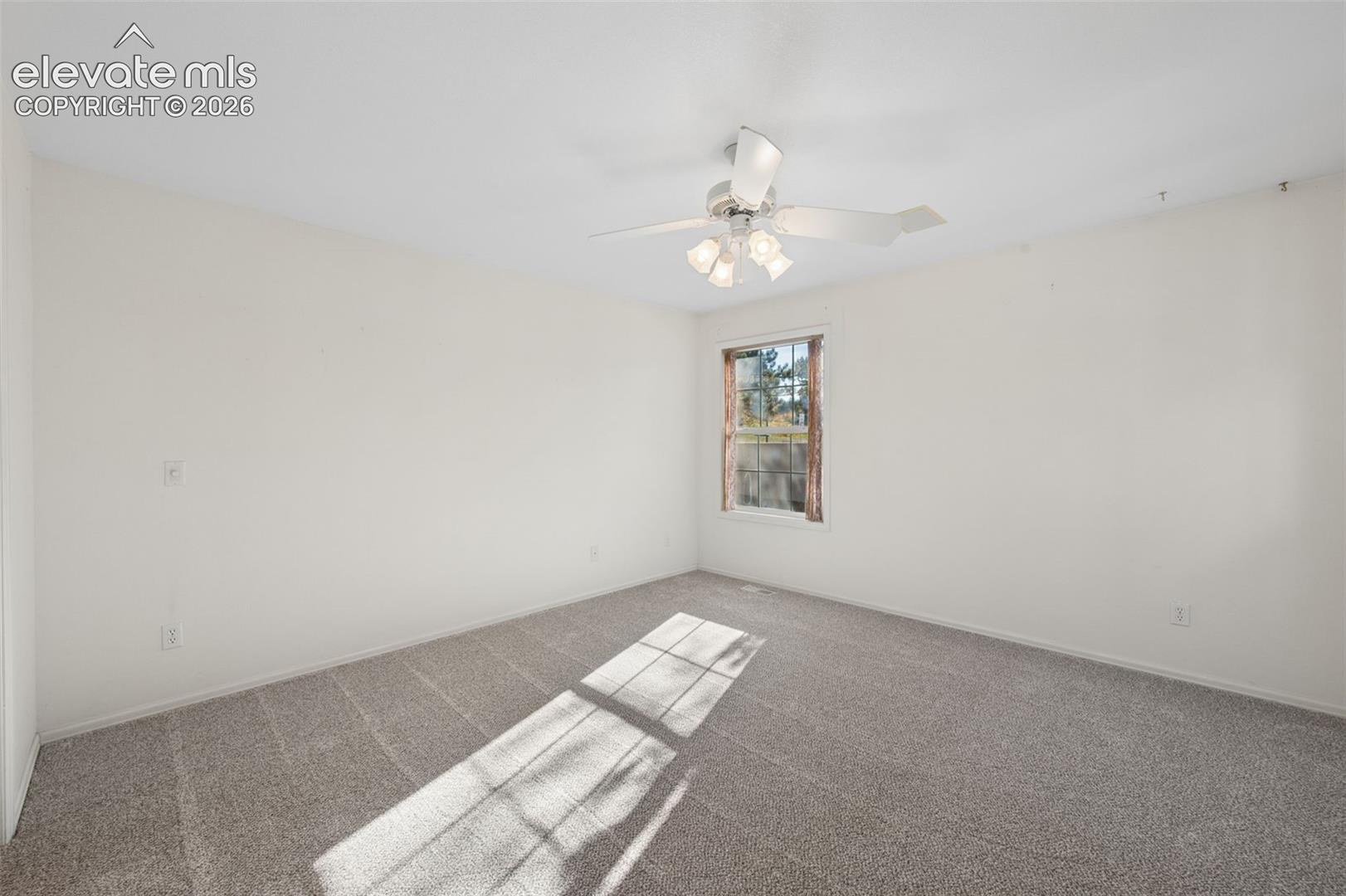 Neutral-toned wall-to-wall carpeting, single-hung window with dark wood-finish trim, and a ceiling-mounted light fixture with a multi-blade fan