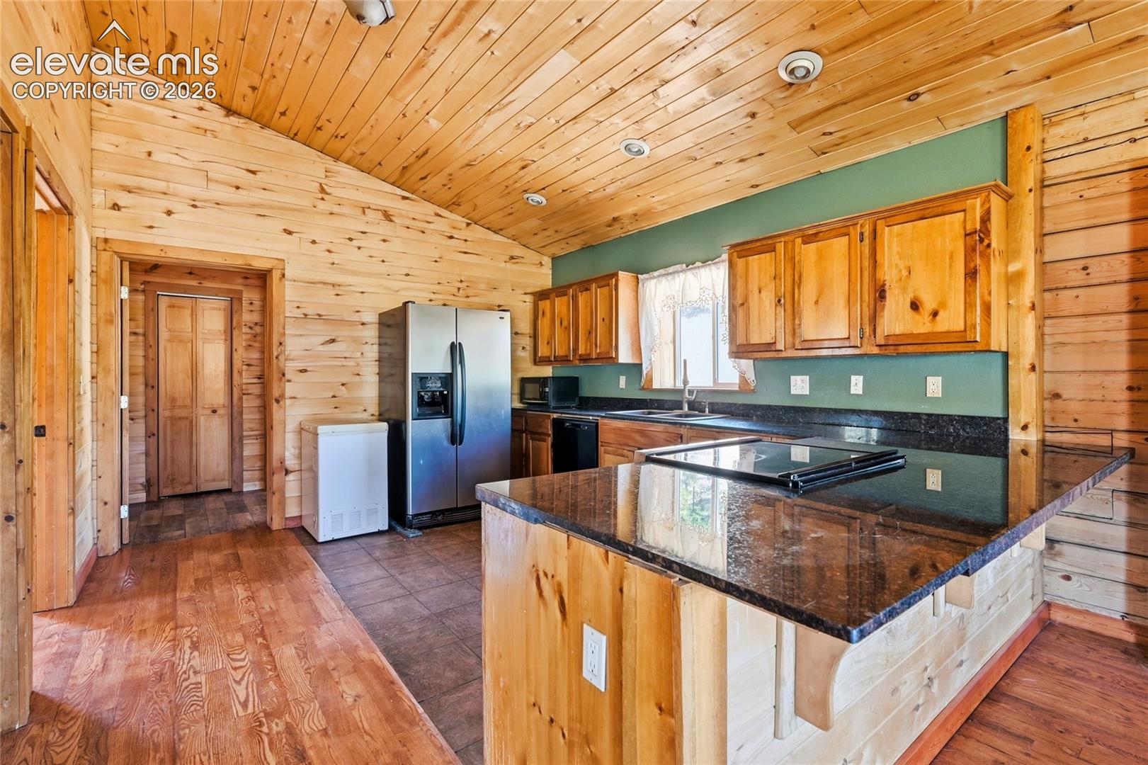 Vaulted wood-paneled ceiling, wood-finish walls, wood cabinetry, granite-finish countertops, and stainless steel refrigerator