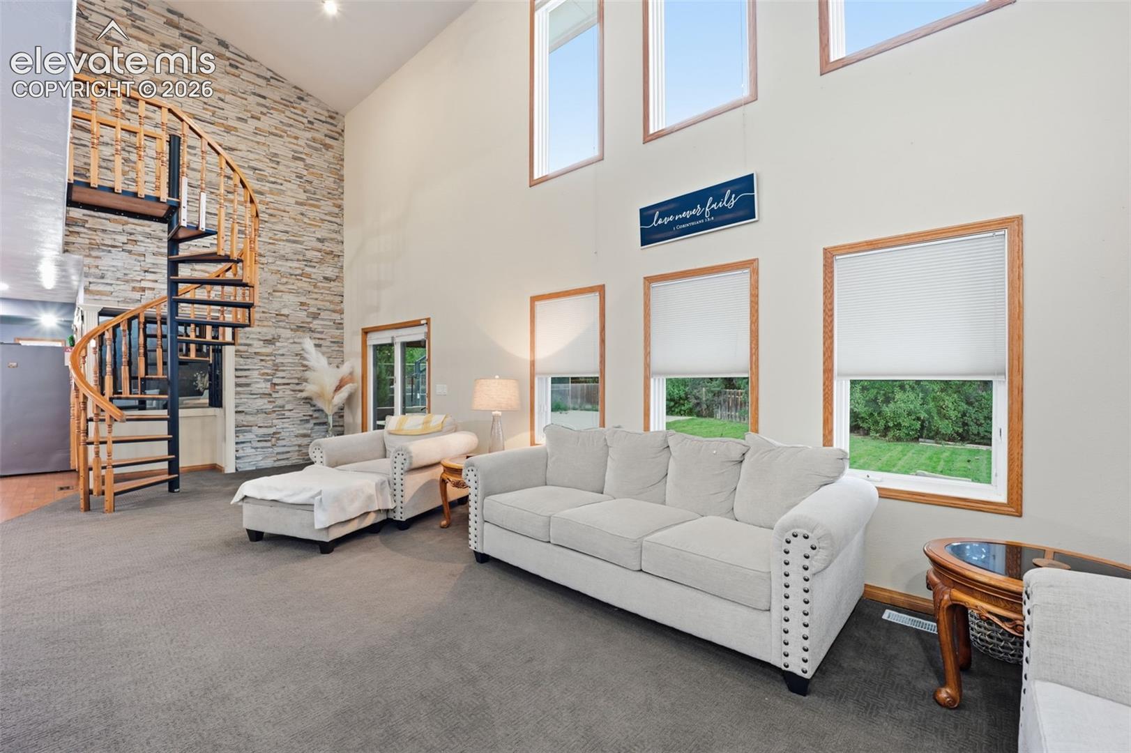 Soaring ceiling with high windows, natural wood-frame windows, stone accent wall, spiral staircase, and gray carpeting