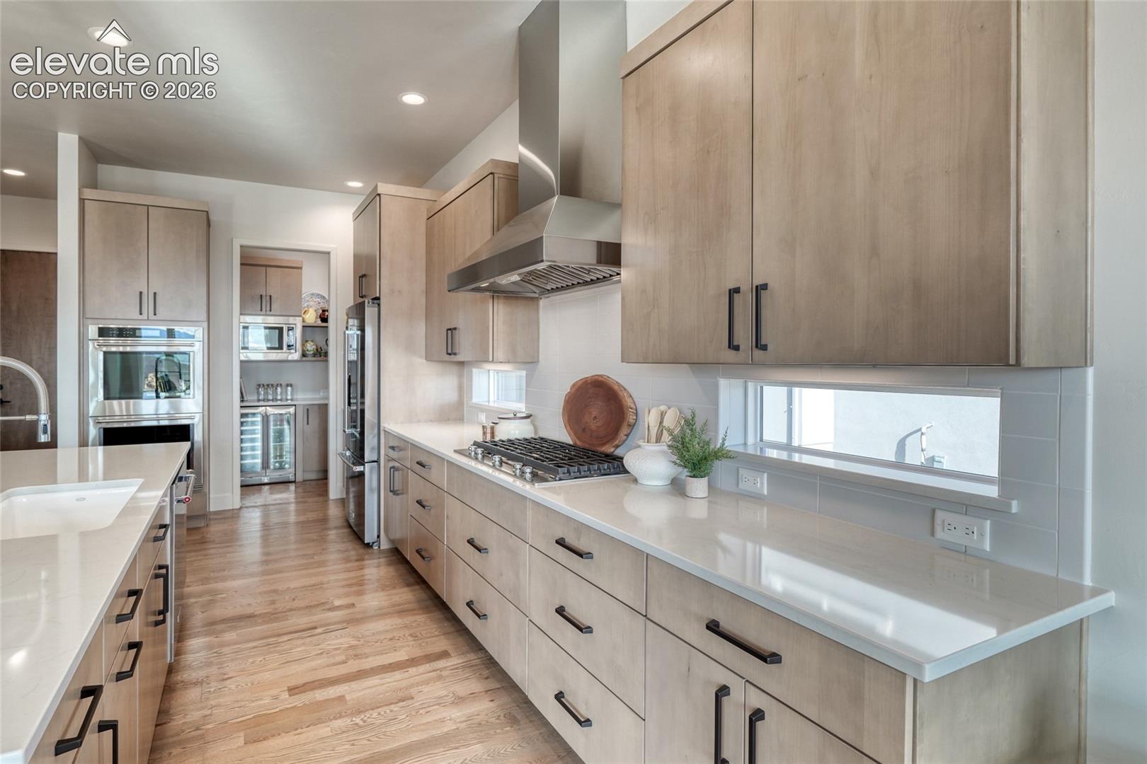 Kitchen featuring custom flat-panel cabinetry, light-toned countertops, stainless steel gas cooktop with chimney-style range hood, built-in double wall ovens, and wood-finish flooring