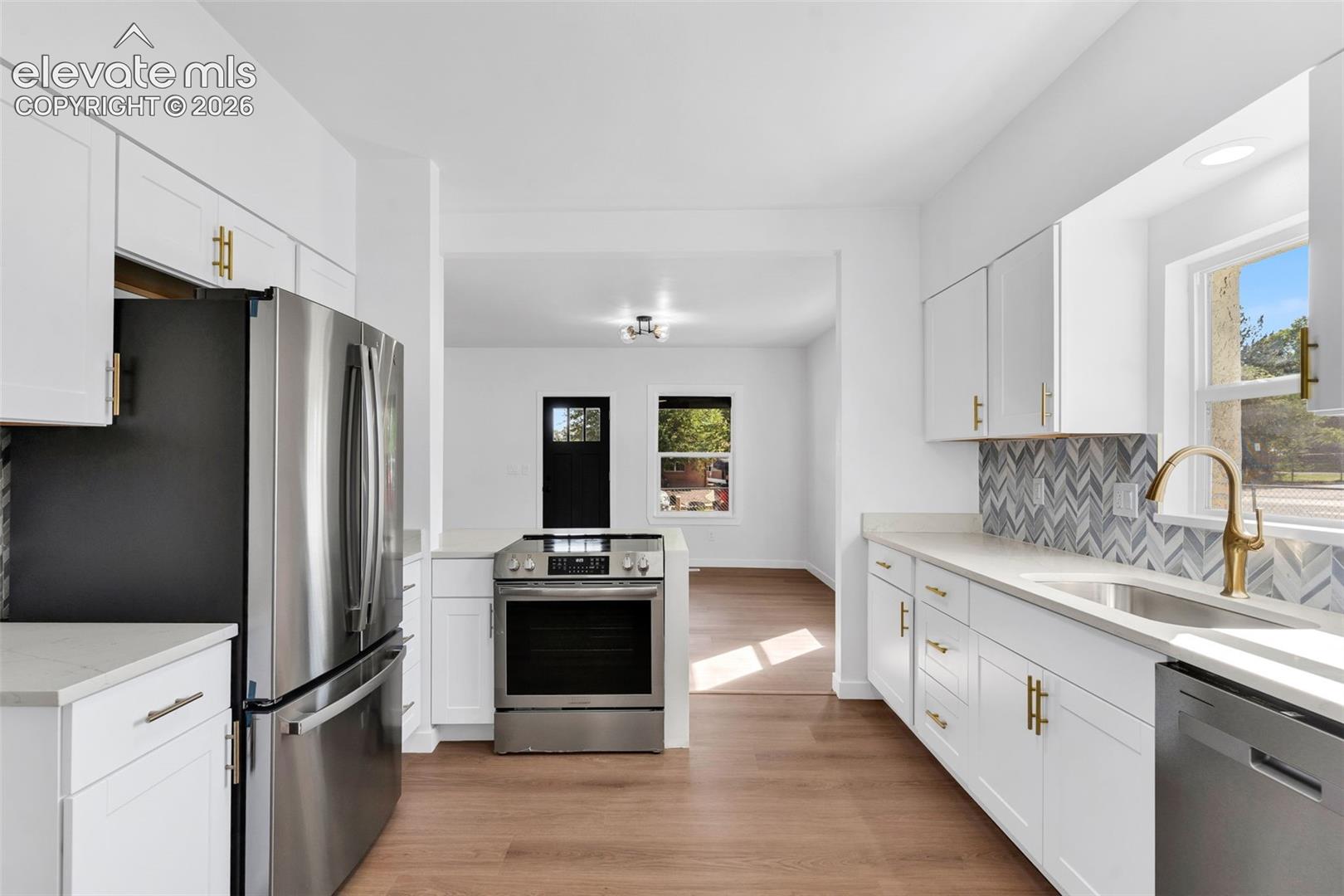 Kitchen featuring white shaker-style cabinetry with gold-tone hardware, stainless steel appliances, herringbone-pattern tile backsplash, solid-surface countertops, and wood-finish flooring