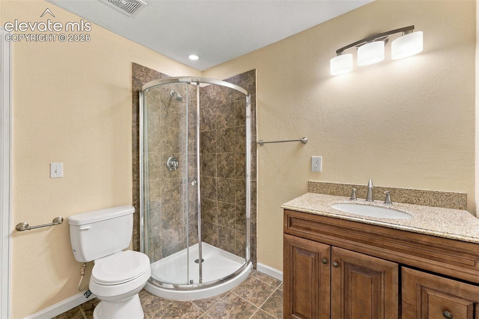 Corner shower with glass enclosure and tiled surround, wood-finish vanity with stone-composite countertop, undermount sink, and three-bulb vanity lighting