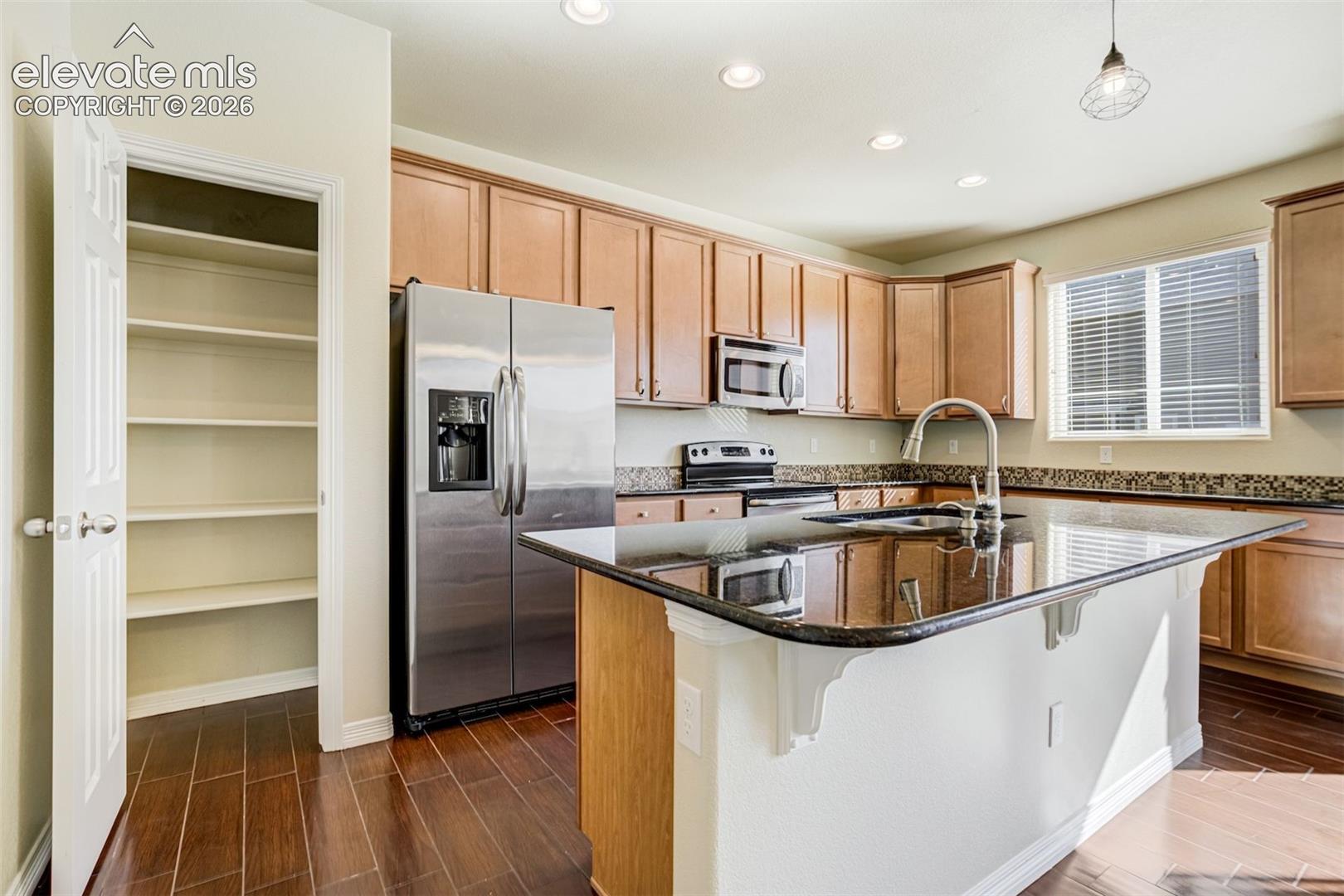 Kitchen featuring wood-finish flooring, recessed lighting, wood cabinetry, built-in stainless steel appliances, and a central island with a sink and pantry