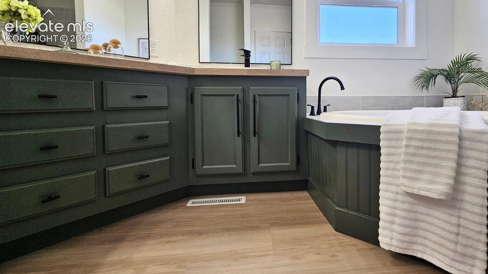 Custom green-painted cabinetry with matte black hardware, light-toned countertop, wood-finish flooring, and a built-in soaking tub featuring a matte black faucet