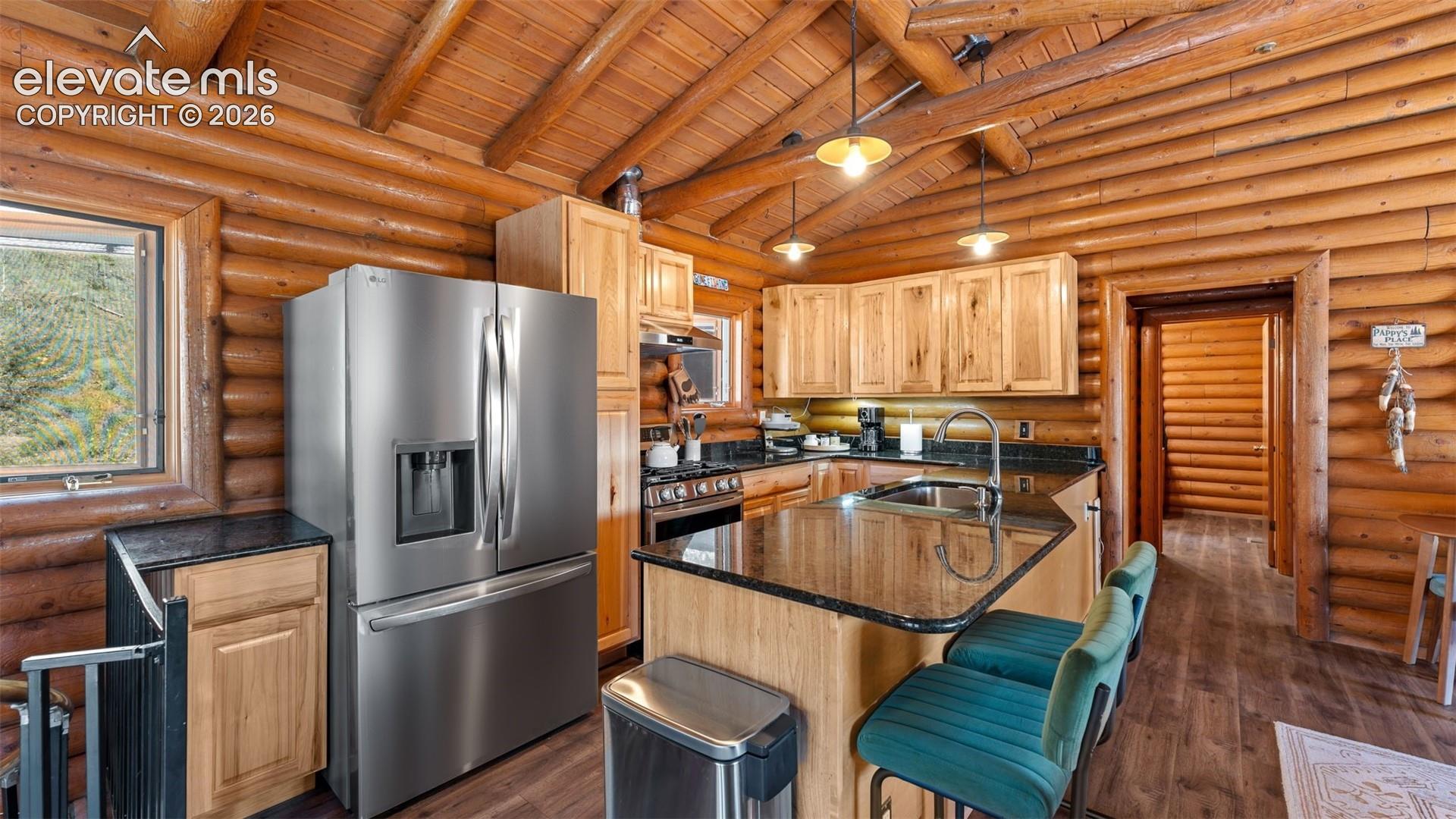 Kitchen featuring exposed log walls, vaulted beam ceiling, stone-finish countertops, stainless steel appliances, and a central island with integrated seating