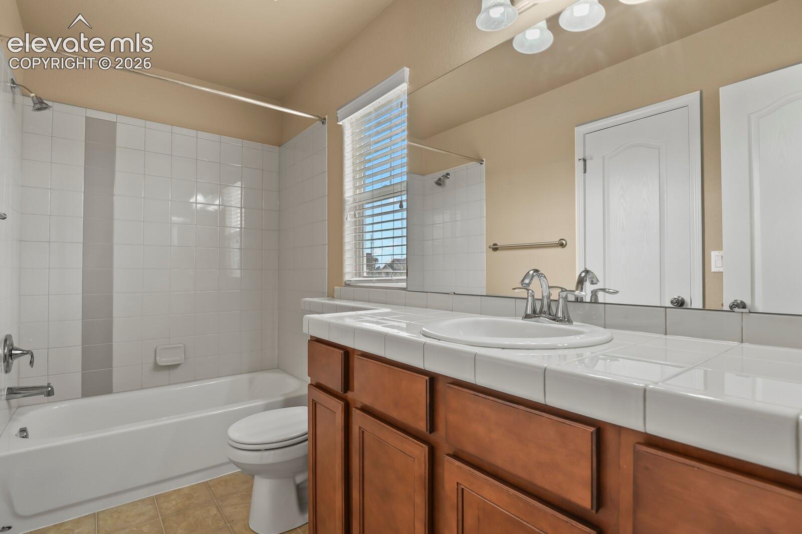 Full vanity mirror, integrated vanity lighting, tile countertop with undermount sink, wood-finish cabinetry, and a shower-tub combination with white tile surround