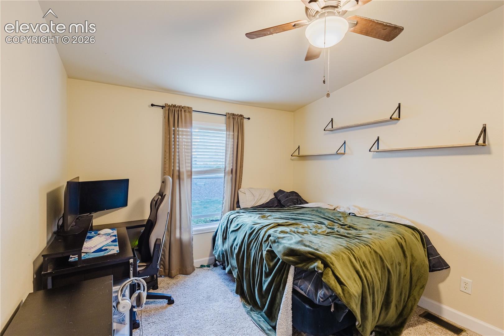 Ceiling fan with light fixture, window with blinds, wall-mounted shelving, and carpeted flooring