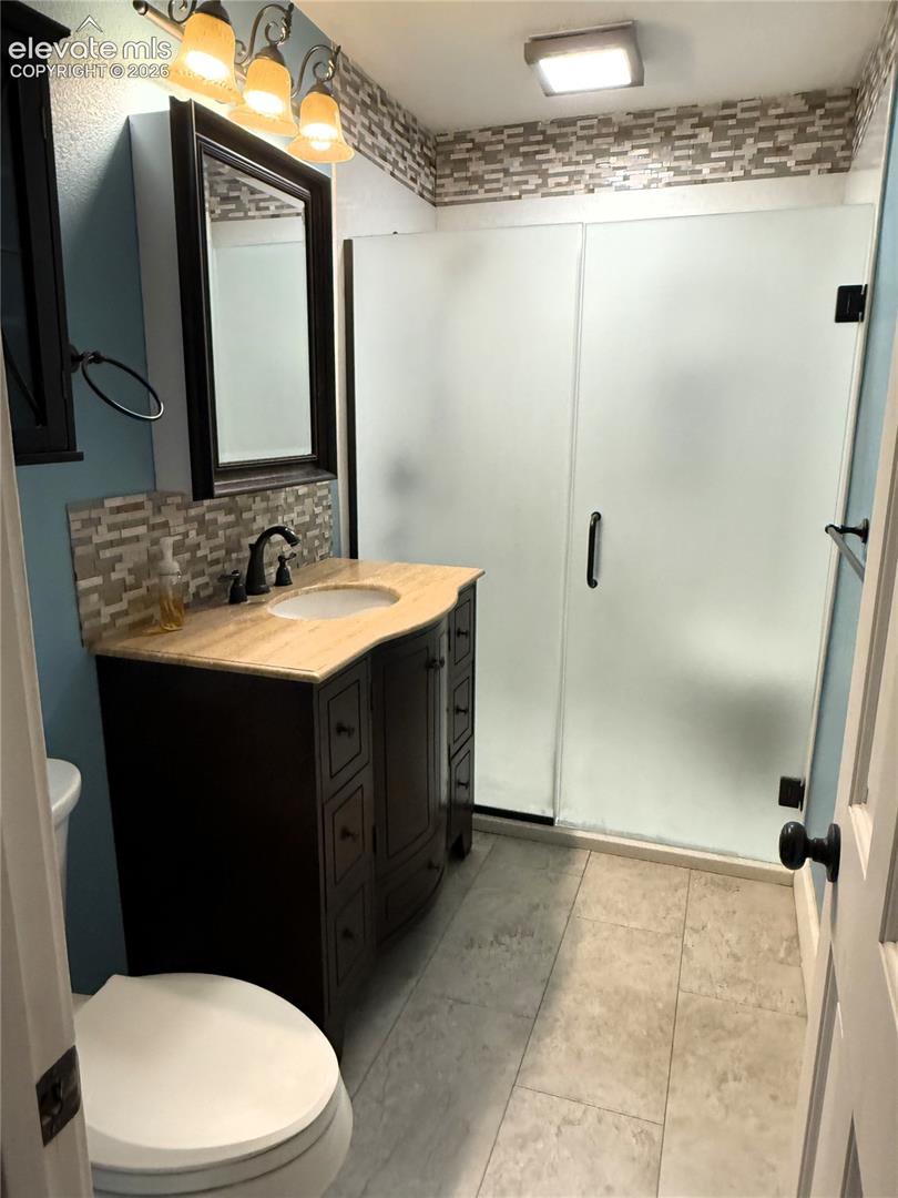Bathroom featuring a dark-finish vanity with a stone-top integrated sink, matte black hardware, a tiled walk-in shower with frosted glass doors, and large-format tile flooring