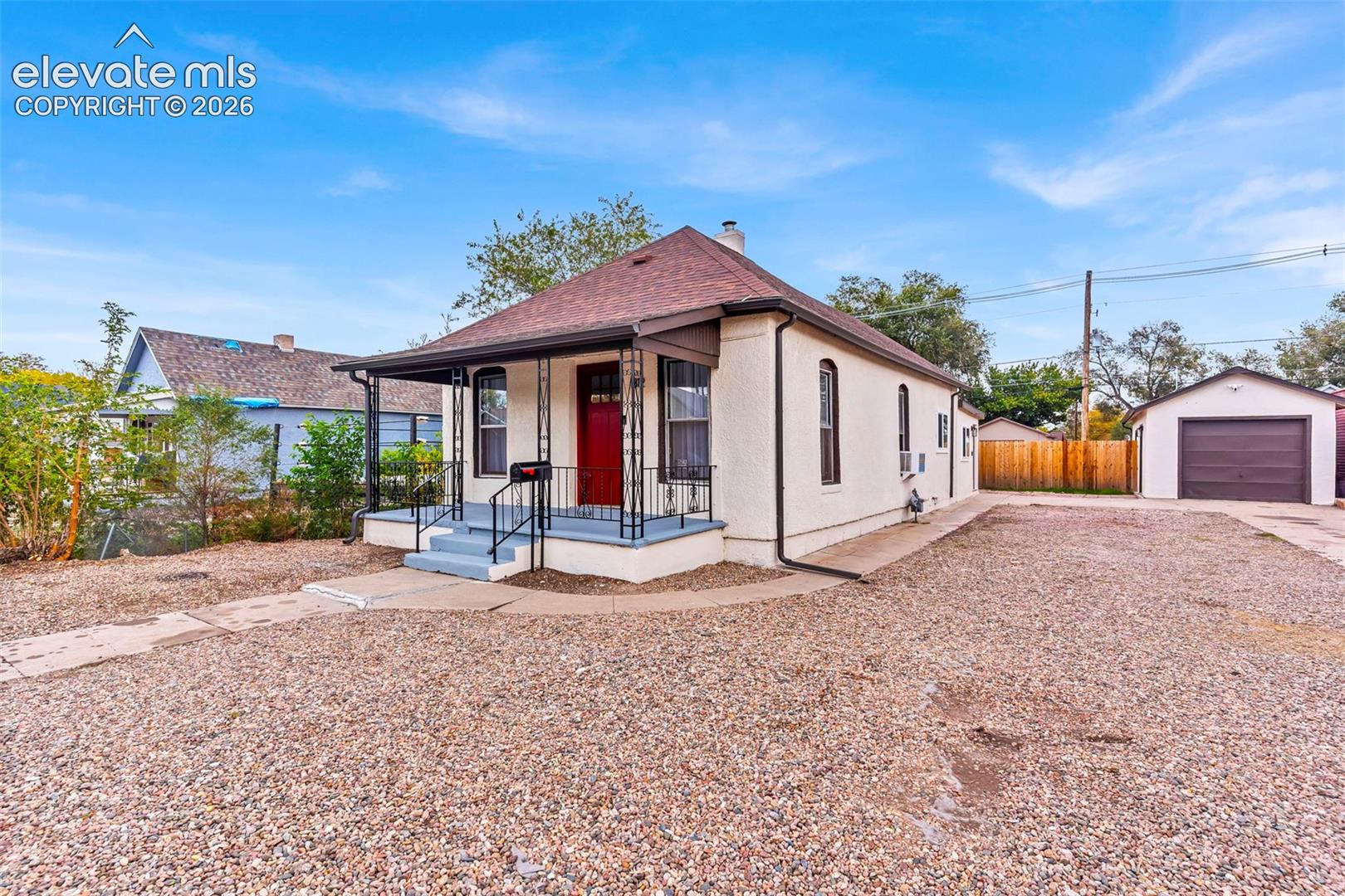 Single-story residence featuring a light-toned exterior, covered front porch with iron railings, multi-panel front door, and a detached garage