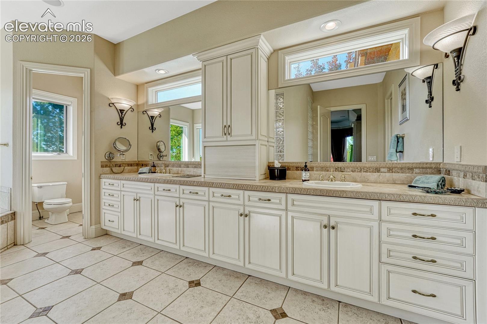Dual-sink vanity with light-tone cabinetry, stone-composite countertops, tile backsplash, transom windows, and tile flooring with decorative inlay