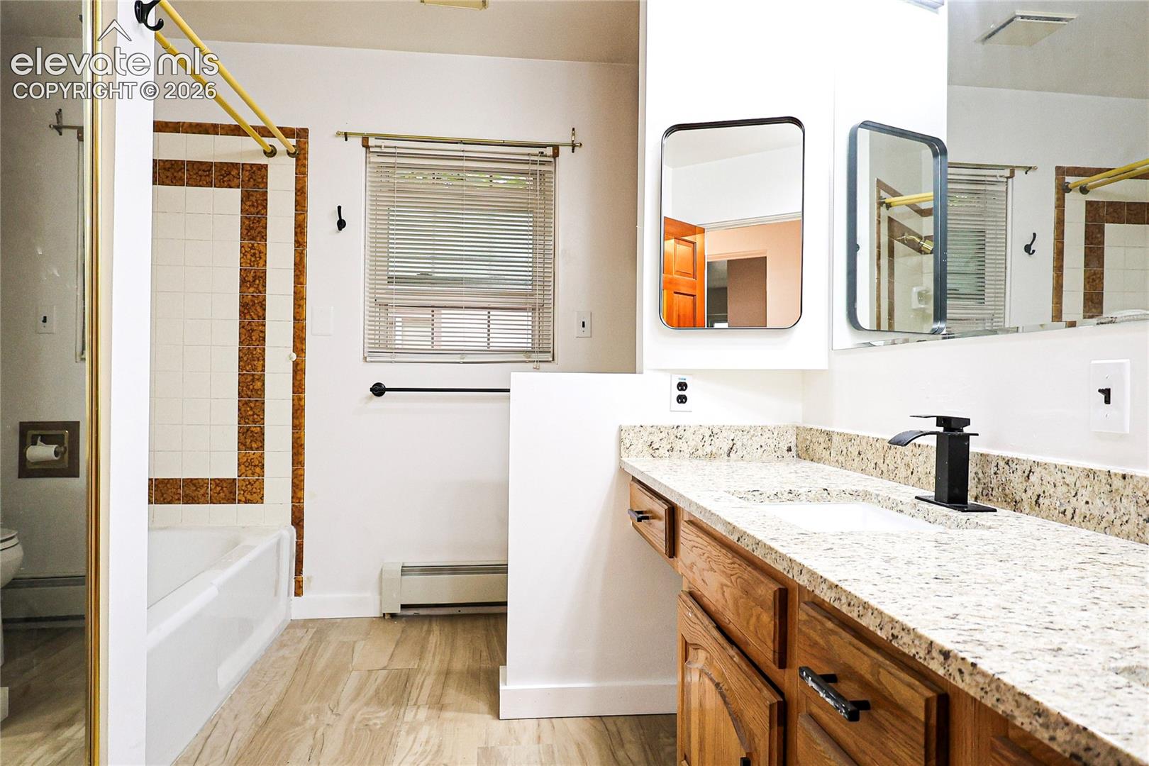Wood-finish flooring, white bathtub with decorative tile surround, functional window, and a vanity with a light-toned countertop and wood cabinetry