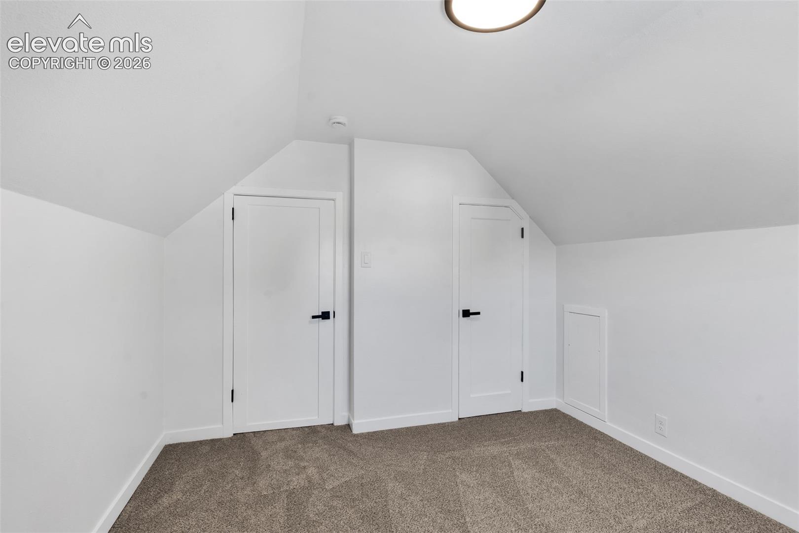 Room featuring vaulted ceilings, neutral-toned carpeting, two shaker-style doors with matte black hardware, and an integrated access panel