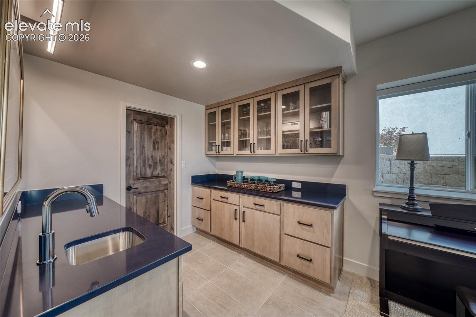 Custom cabinetry with glass-front uppers, solid-surface countertops, integrated sink with gooseneck faucet, tile flooring, and a textured wood-grain interior door