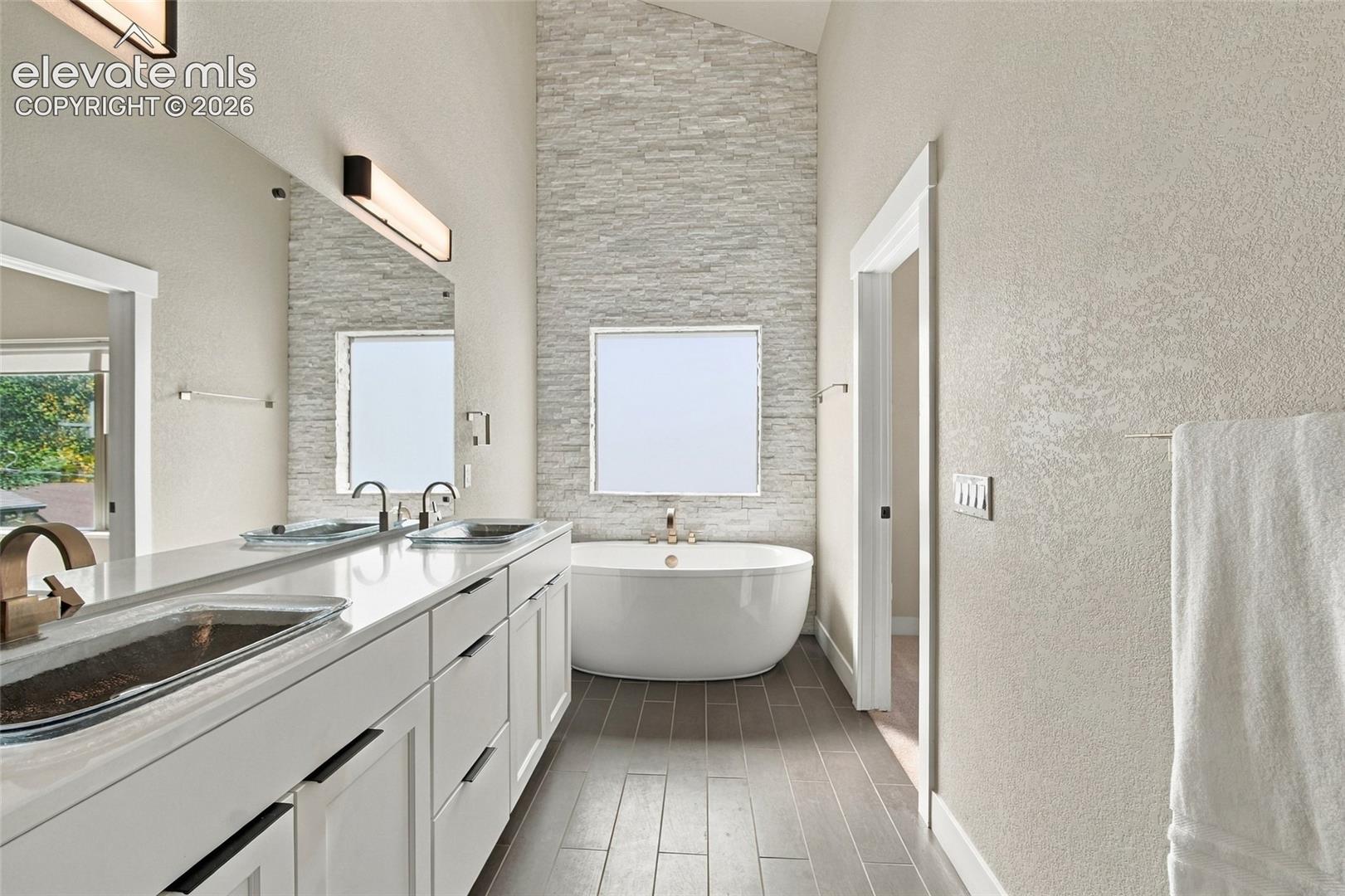 Freestanding soaking tub set against a floor-to-ceiling stacked stone accent wall, double vanity with dual sinks, modern linear vanity lighting