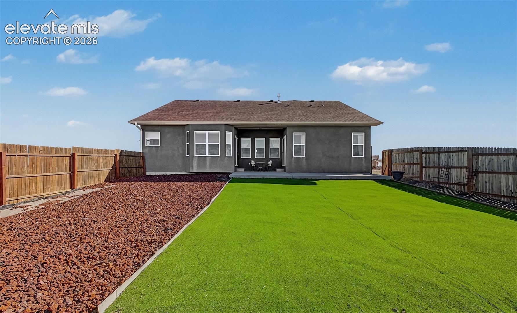 Rear exterior featuring synthetic turf, decorative stone landscaping, a concrete patio, and perimeter wood fencing