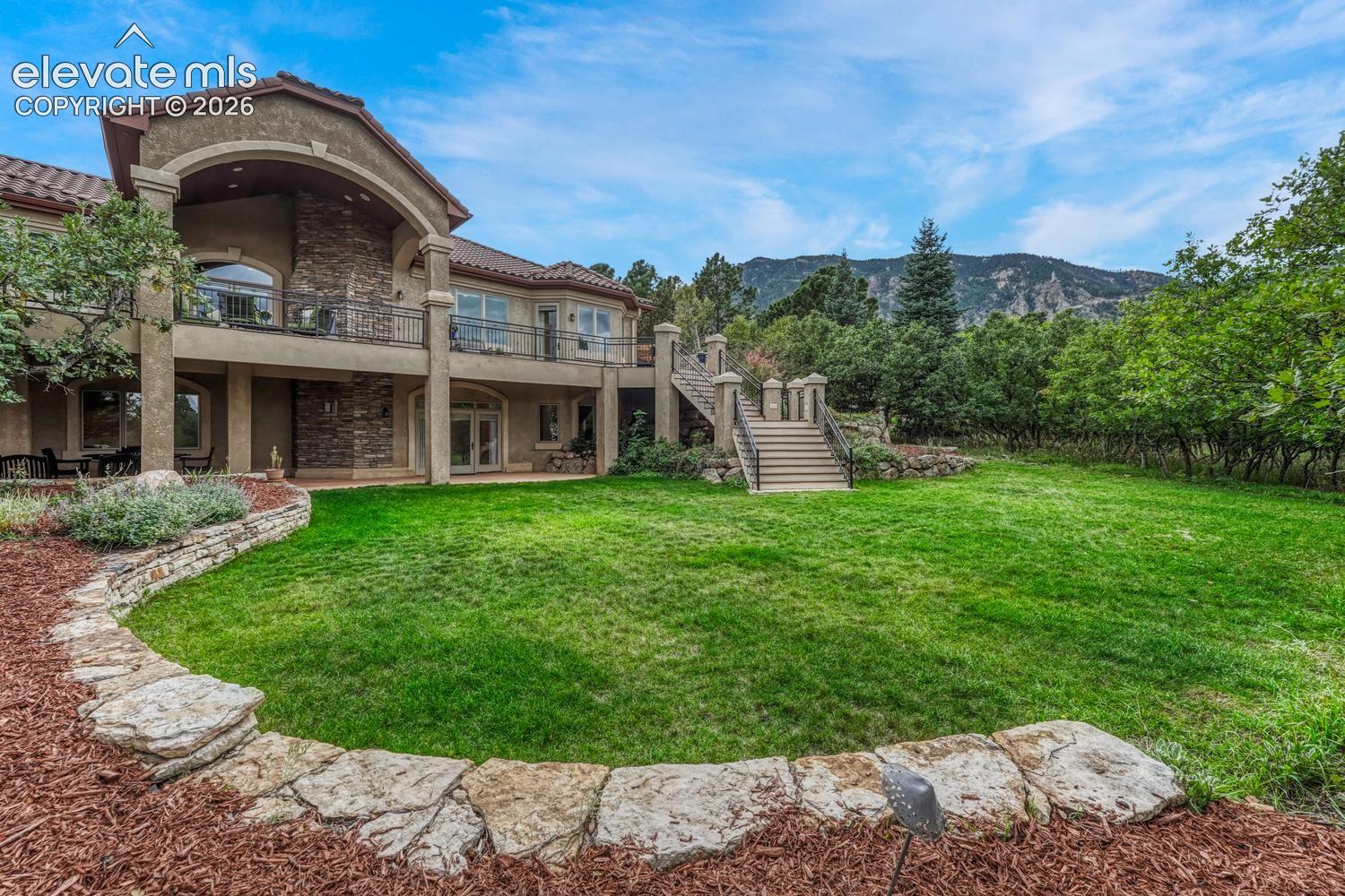 Stucco exterior featuring stacked stone masonry accents, a wrap-around balcony with iron railings, an external staircase, and a tiered stone-retaining wall surrounding the lawn