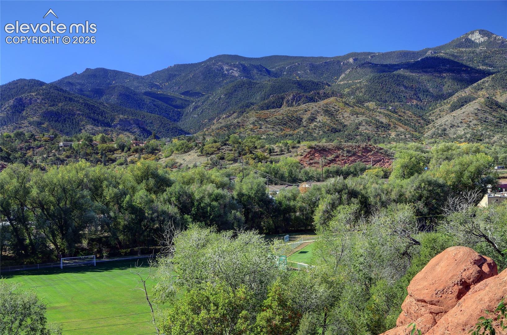 Expansive mountain backdrop featuring prominent geological rock formations, mature deciduous trees, and a maintained green grass field