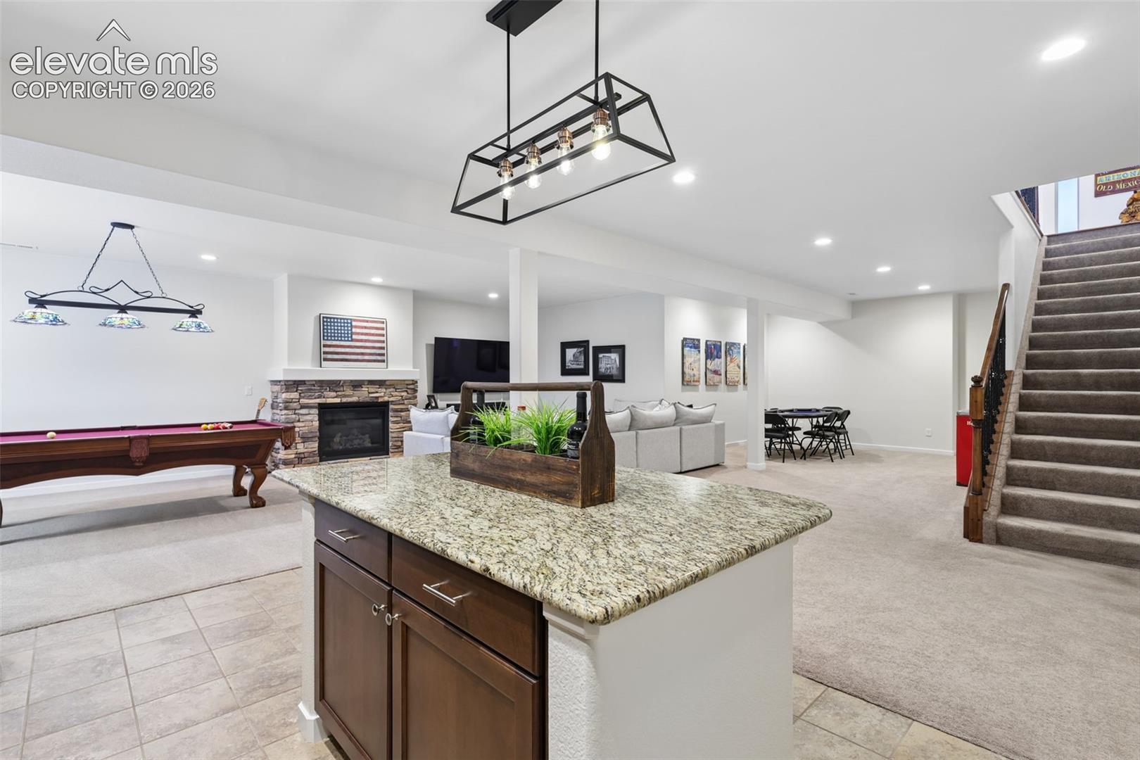 Open-concept lower level with recessed lighting, a stone-faced fireplace, and a wood-finish staircase with dark iron balusters