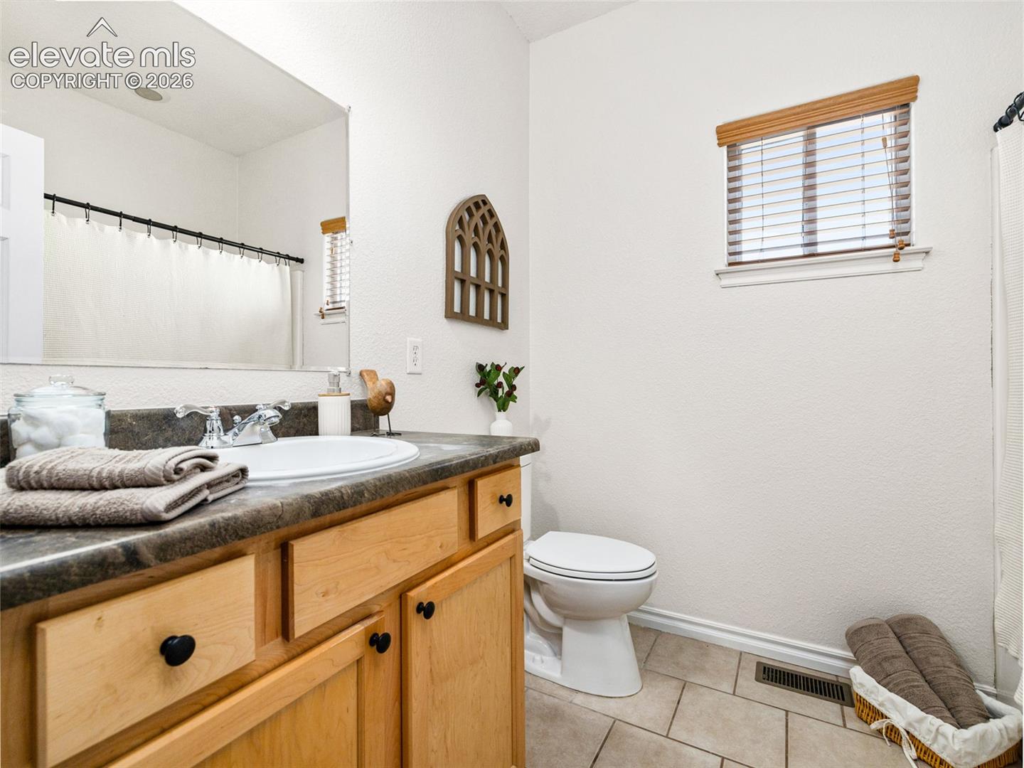 Vanity with natural wood cabinetry and dark countertop, integrated sink with chrome faucet, large wall mirror, and tiled flooring