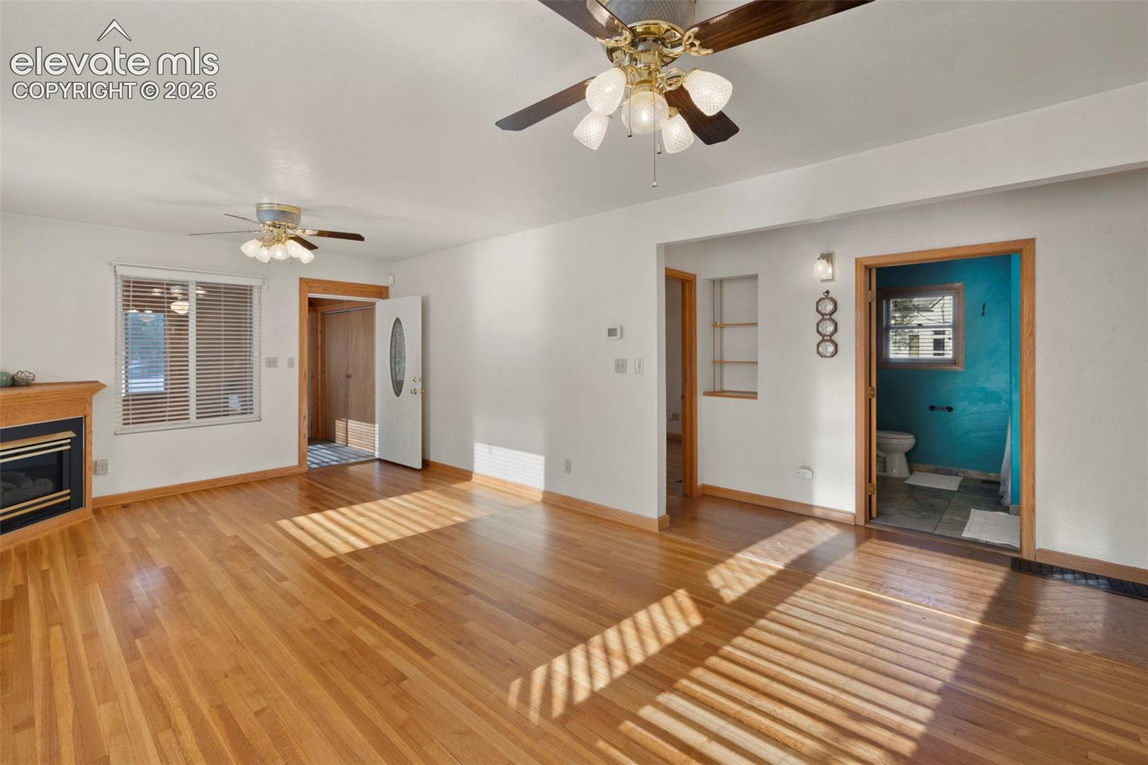 Hardwood flooring throughout, two ceiling fans with integrated lighting, a fireplace with a wood-finish mantel, and white window blinds