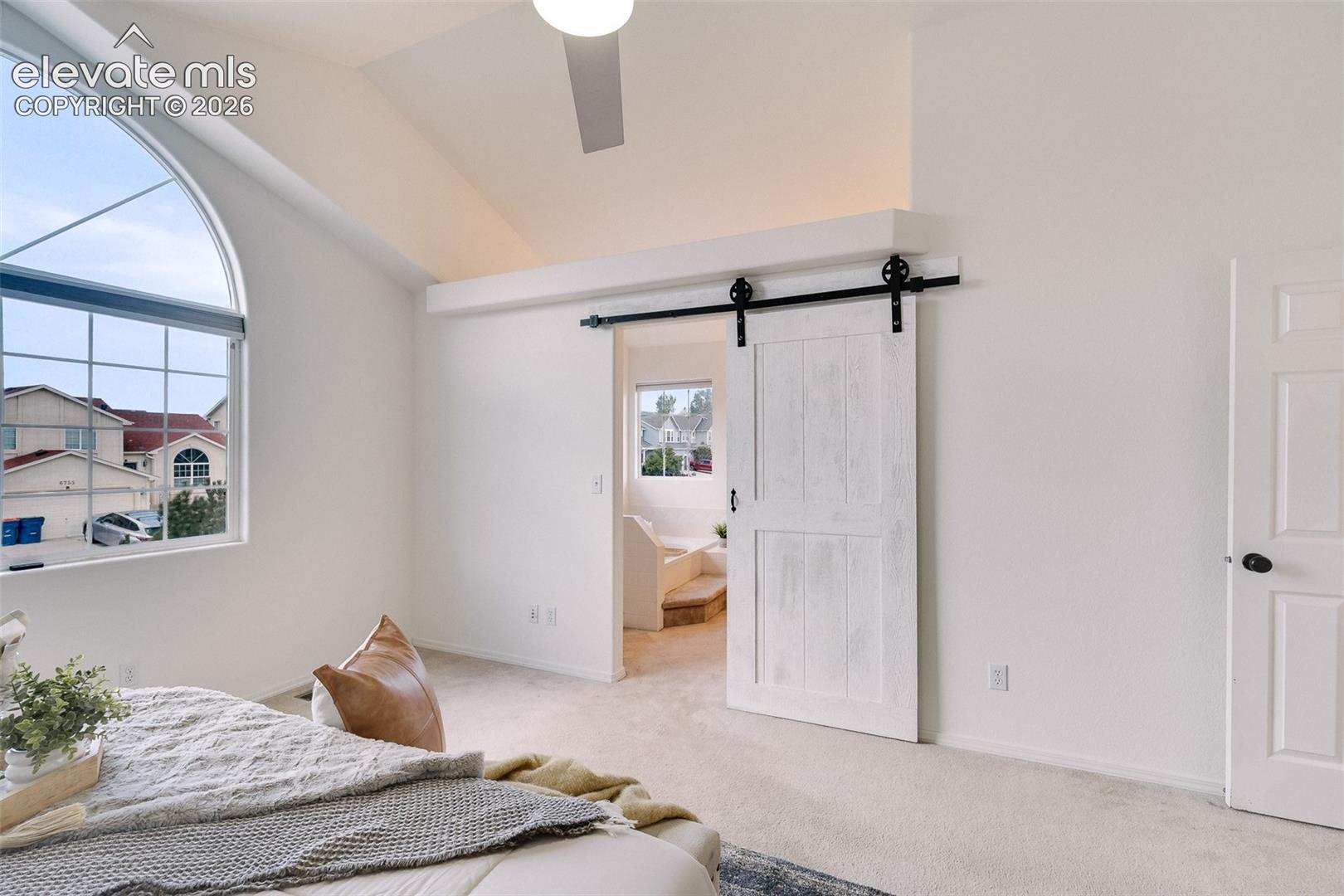 Arched window with gridded panes, vaulted ceiling, ceiling fan, white barn door, and light-colored carpeting