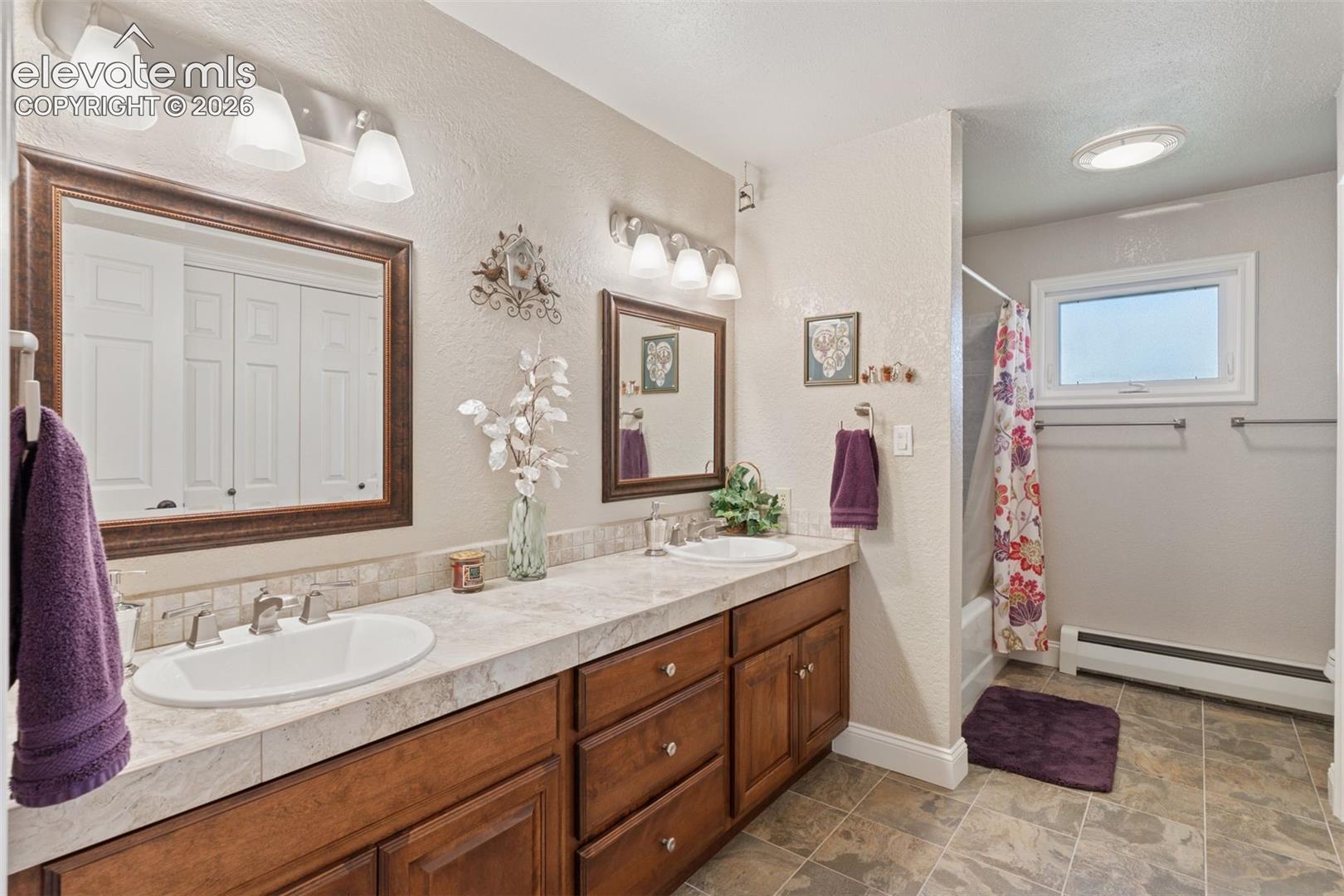 Additional full bathroom with double vanity, shower / bath combo, a textured wall, and stone flooring that connects multiple ground level bedrooms