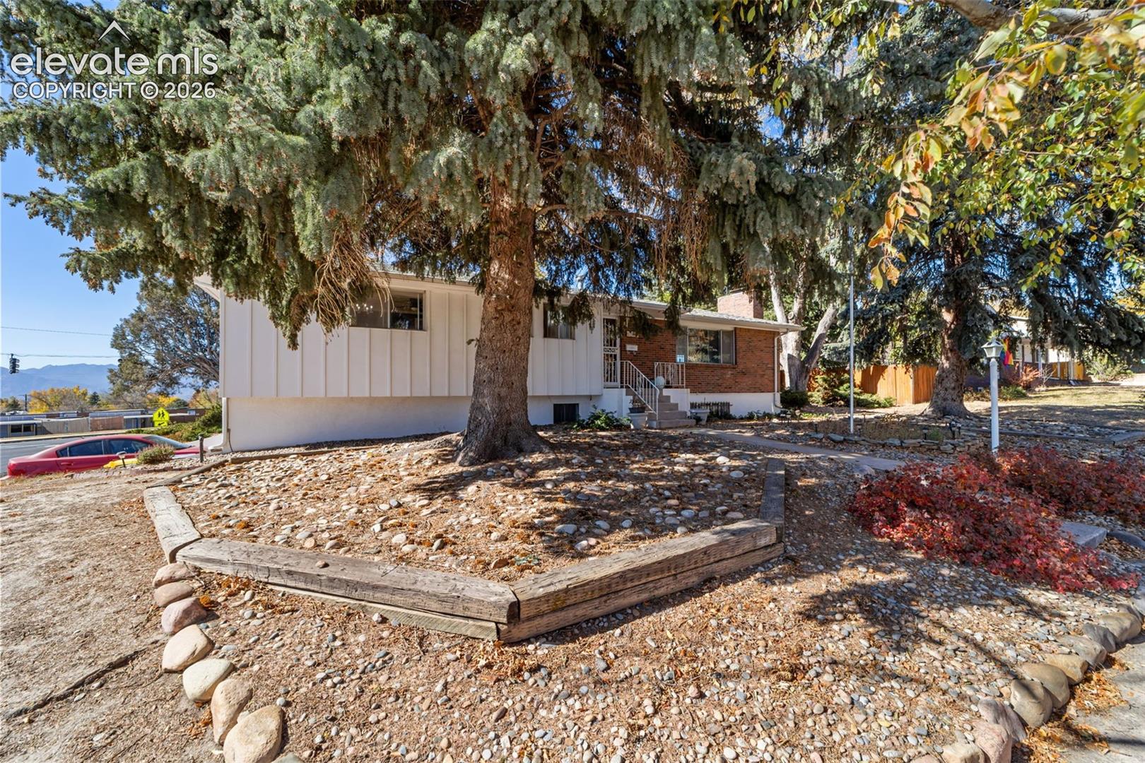 Split-level residence featuring a brick and vertical siding exterior, mature evergreen trees, and a terraced front yard with timber retaining walls