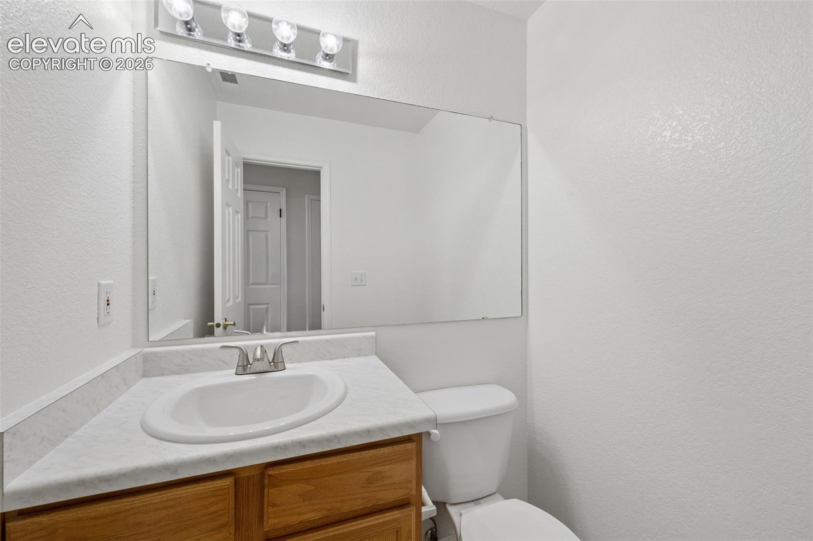 Wood-finish vanity cabinet, solid-surface countertop with integrated basin, polished metal faucet, large wall-mounted mirror, and four-light vanity fixture