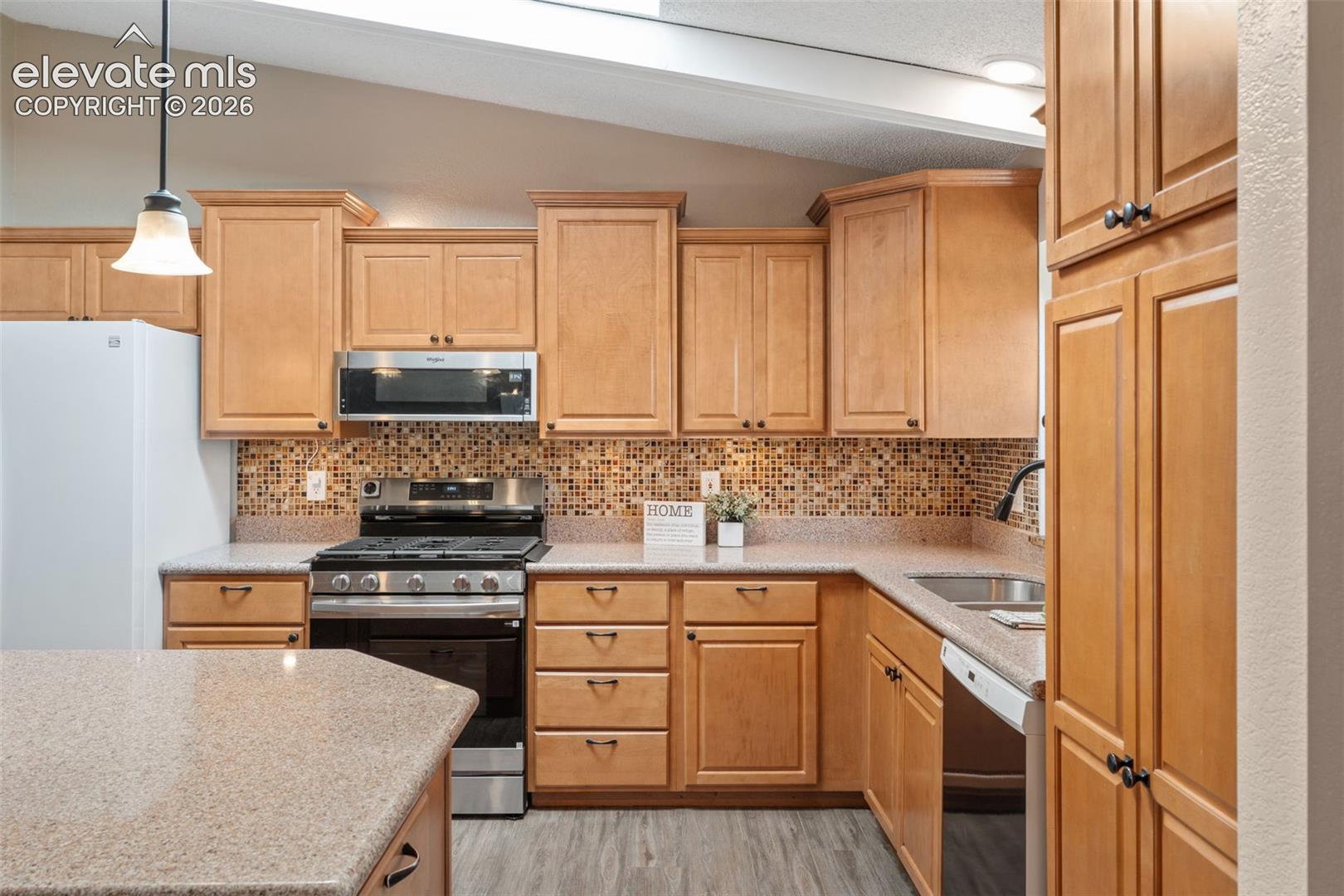 Kitchen featuring natural wood cabinetry, stone-texture countertops, mosaic tile backsplash, stainless steel appliances, and a vaulted ceiling with skylight
