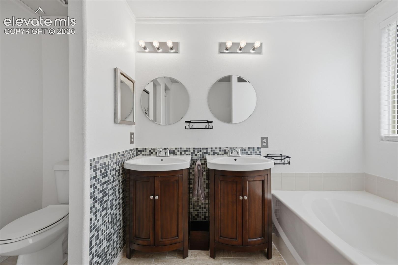 Dual vanities with wood-finish cabinetry, mosaic tile wainscoting, and individual circular mirrors