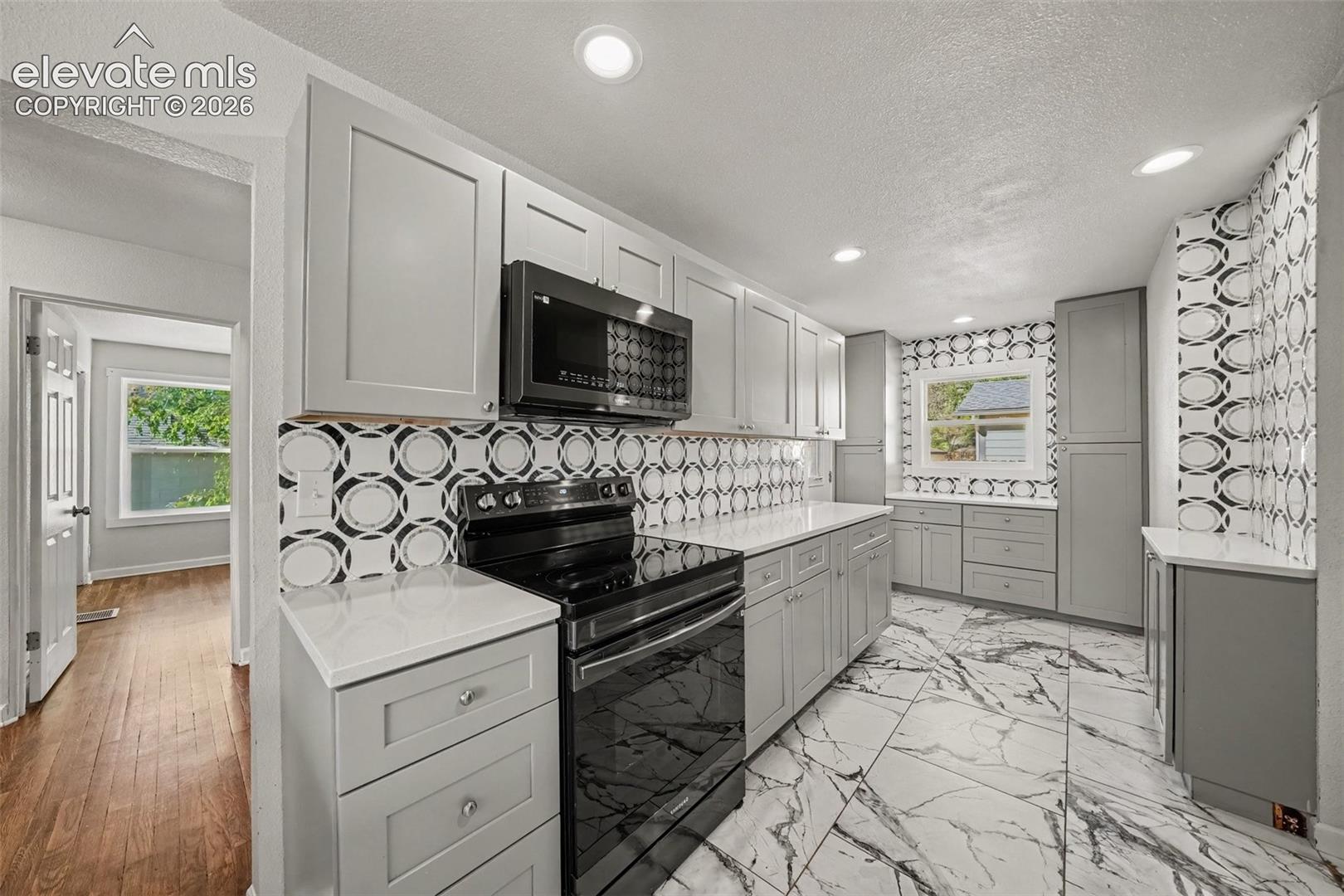 Kitchen featuring grey shaker-style cabinetry, solid-surface countertops, patterned tile backsplash, large-format veined floor tile, and built-in black appliances