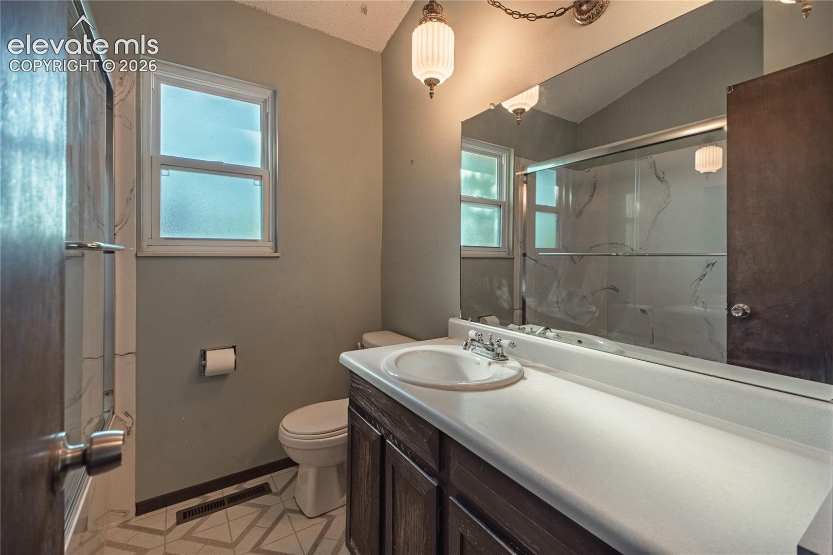 Bathroom featuring a dark wood vanity with a white countertop, oval undermount sink, large wall-to-wall mirror, glass-enclosed shower, and geometric patterned flooring
