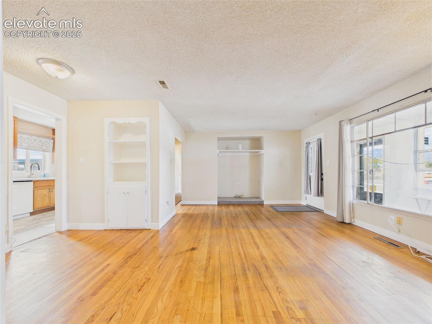 Expansive view of the main living area showcasing hardwood flooring, a large front window, built-in cabinetry, and an open connection to the kitchen.